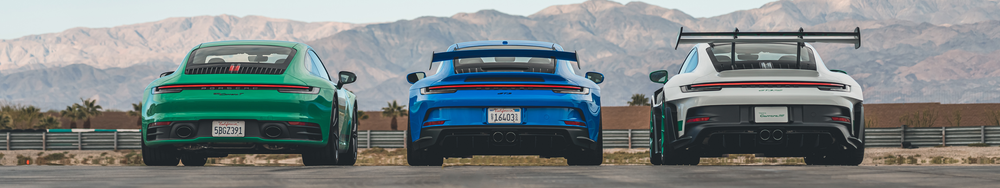 Rear view of three Porsche GT cars in green, blue, and white, parked on a race track with mountains in the background.