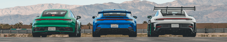 Rear view of three Porsche GT cars in green, blue, and white, parked on a race track with mountains in the background.