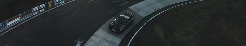 A black sports car, likely a Porsche, on a race track, viewed from above in a bird's eye perspective, with curbing painted in white, red, and blue on the left side.