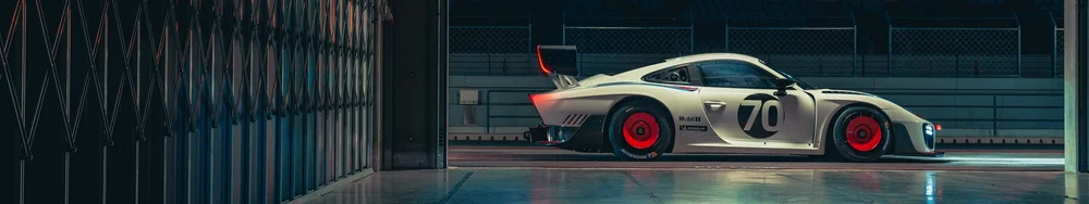 A white race car with the number 70 on its side and red wheels is parked in a garage or pit lane area at night, with fencing and a lit track in the background.