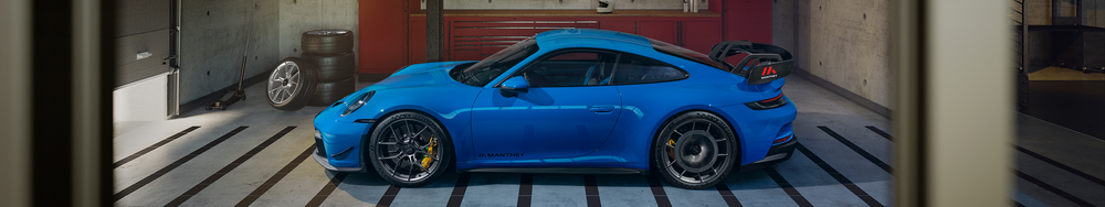 A blue sports car parked in a garage. The garage has black and white striped lines on the floor and red cabinets in the background. The vehicle has a rear wing, black wheels, and yellow brake calipers.
