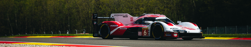A red and white Porsche 963 race car driving on a racetrack with grass and trees in the background.