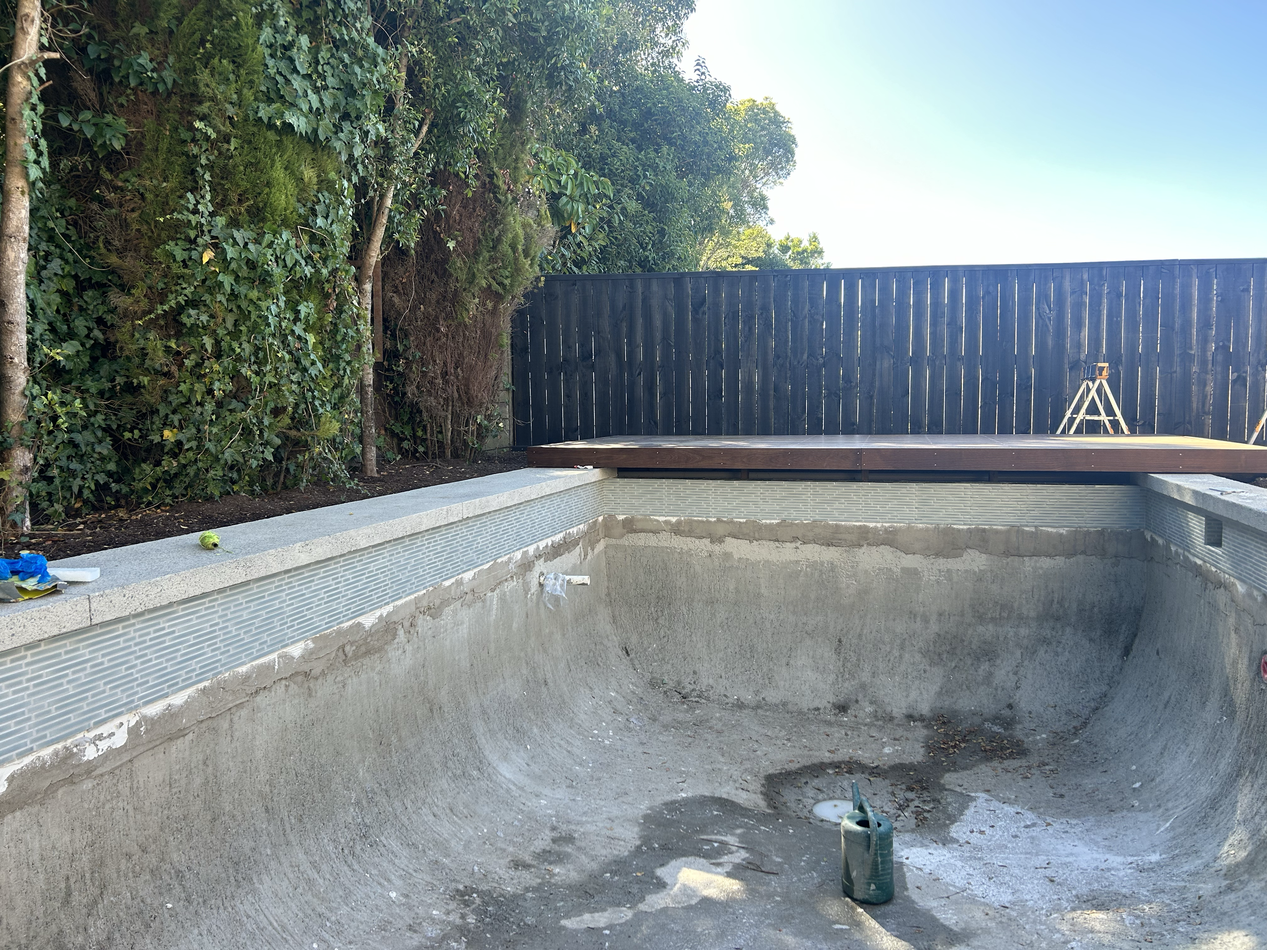 Empty swimming pool under renovation with a watering can inside, surrounded by construction tools and debris, with green bushes and trees behind a black wooden fence. Existing pool refurbishment Built by ESC outdoor living swimming pool builders. Auc