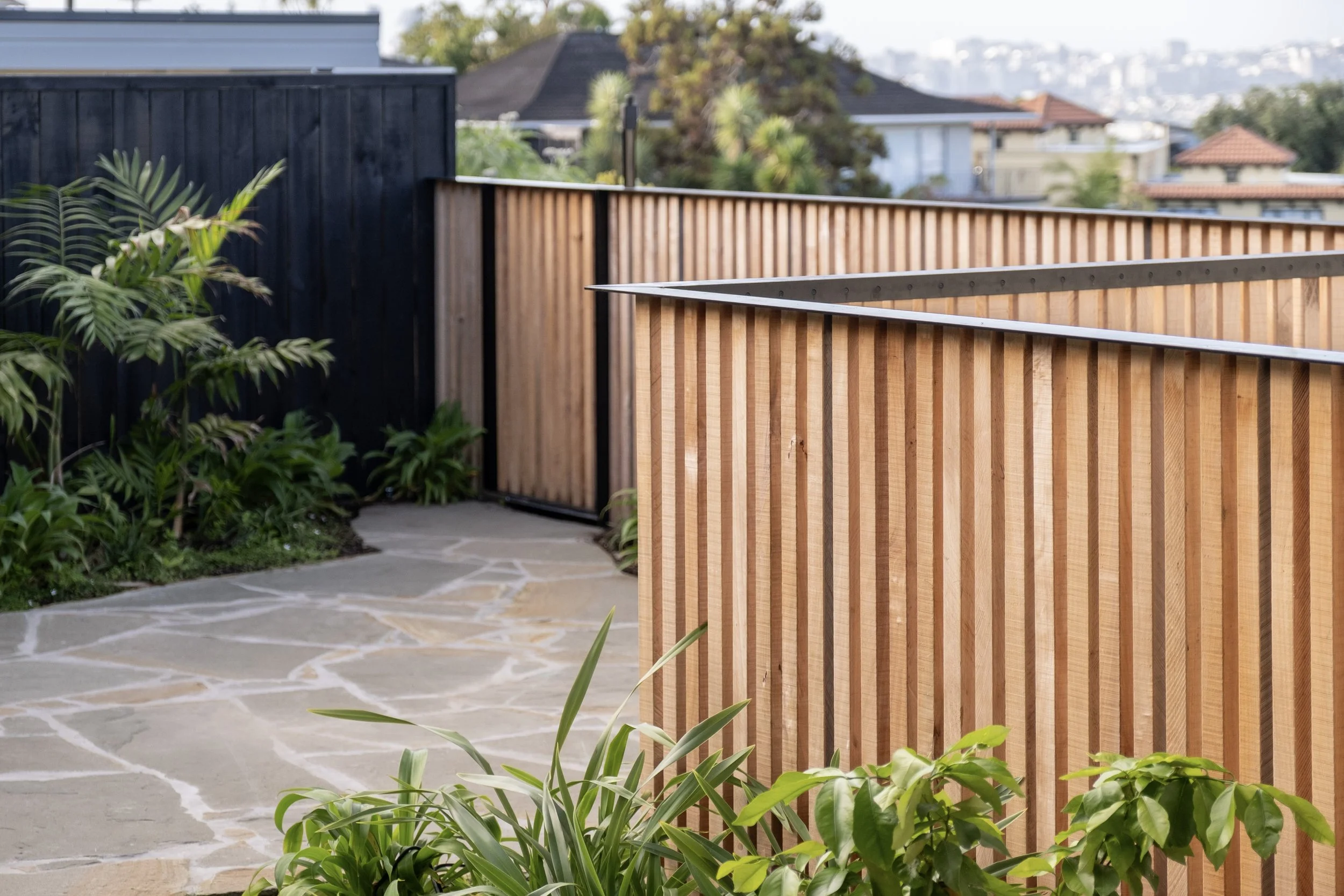 A backyard with a stone pathway, lush green plants, and a wooden fence with vertical slats. Existing pool refurbishment Built by ESC outdoor living swimming pool builders. Auckland. skilled builders