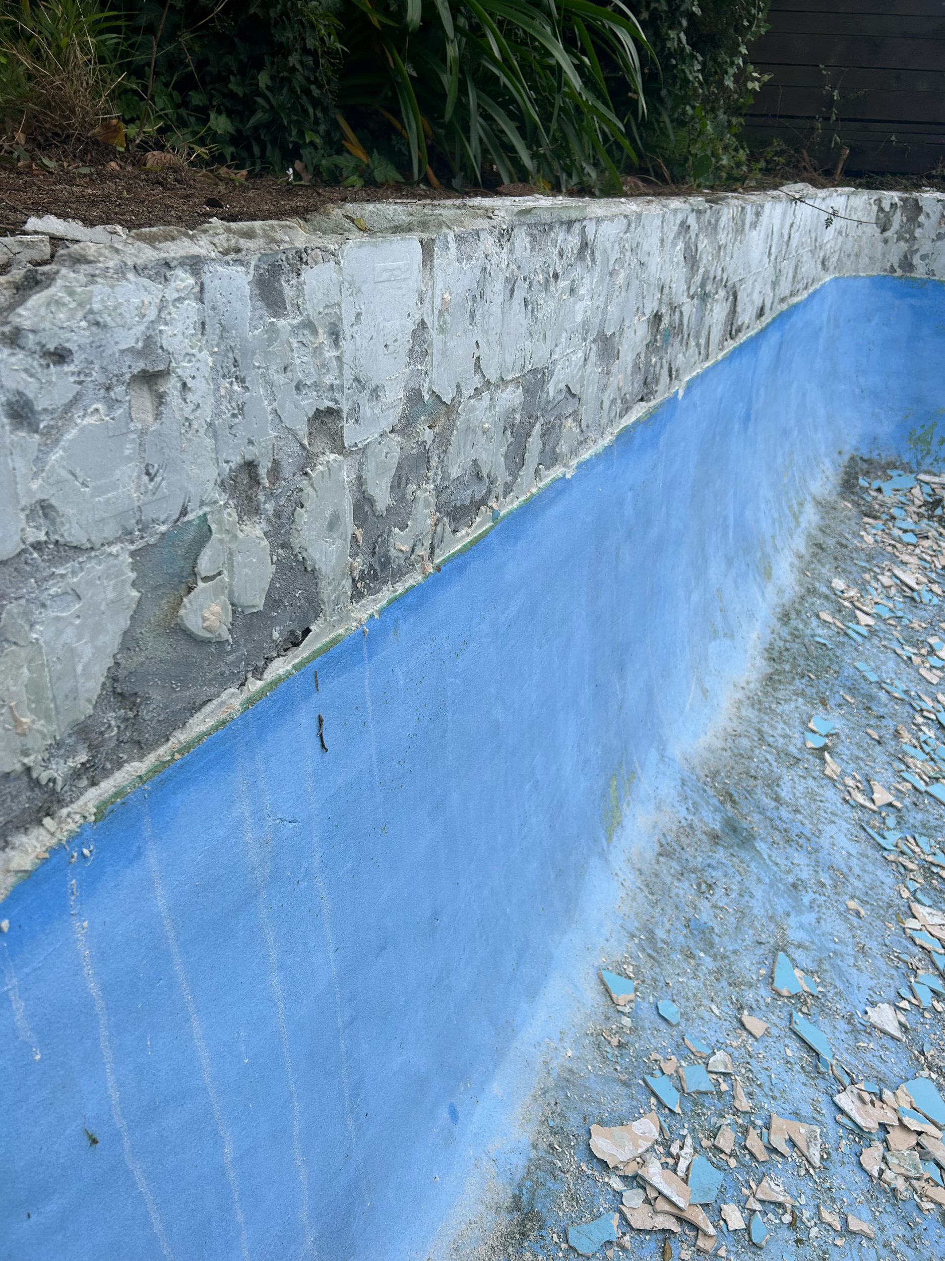 Close-up of the edge of a swimming pool with a blue interior surface and a stone retaining wall above it, surrounded by dirt and broken tiles. Existing pool refurbishment Built by ESC outdoor living swimming pool builders. Auckland.