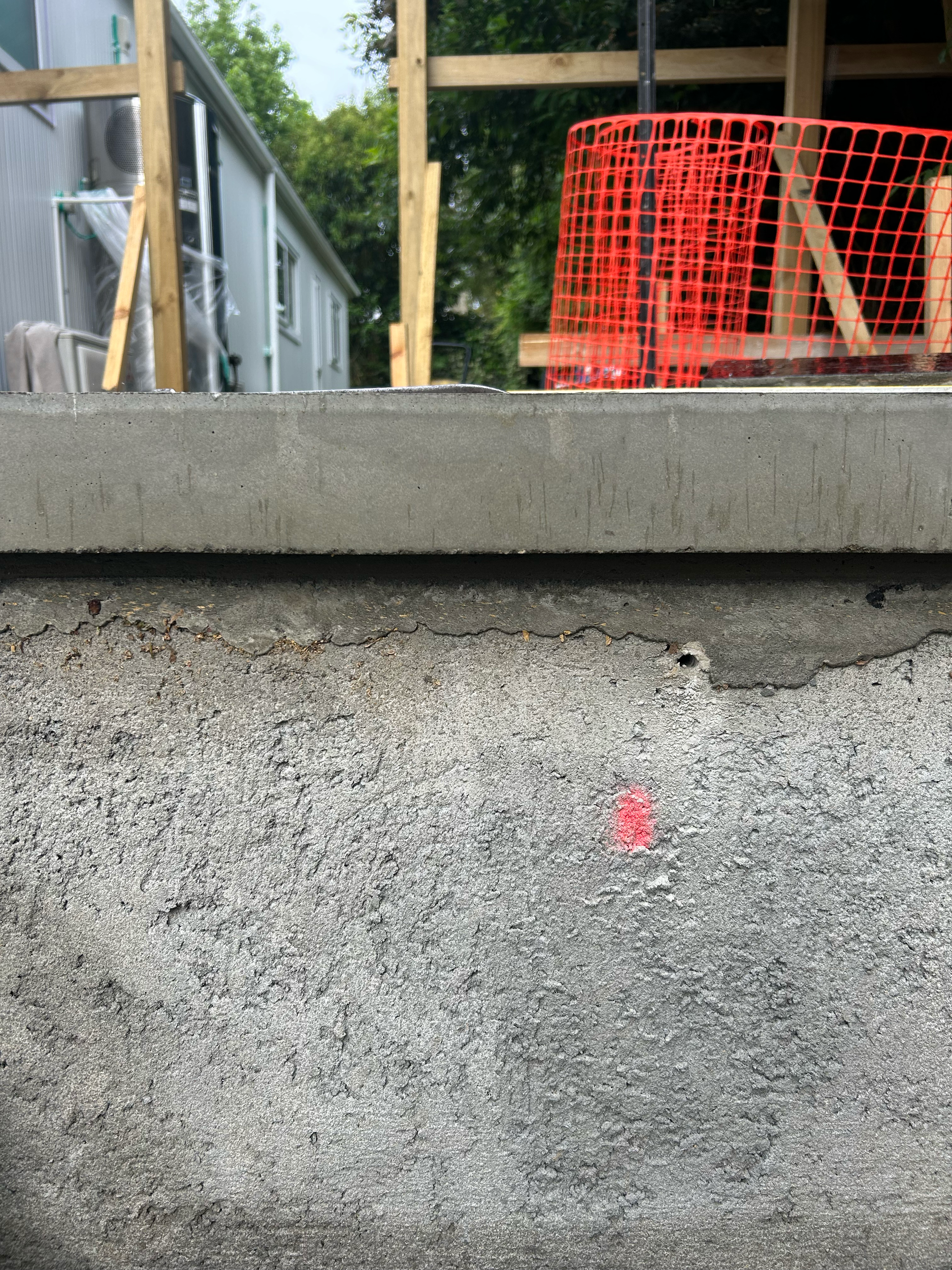 Close-up of a concrete wall with a small pink spray paint dot, with a construction site in the background showing wooden framing, a red safety barrier, and building materials. Existing pool refurbishment Built by ESC outdoor living swimming pool buil