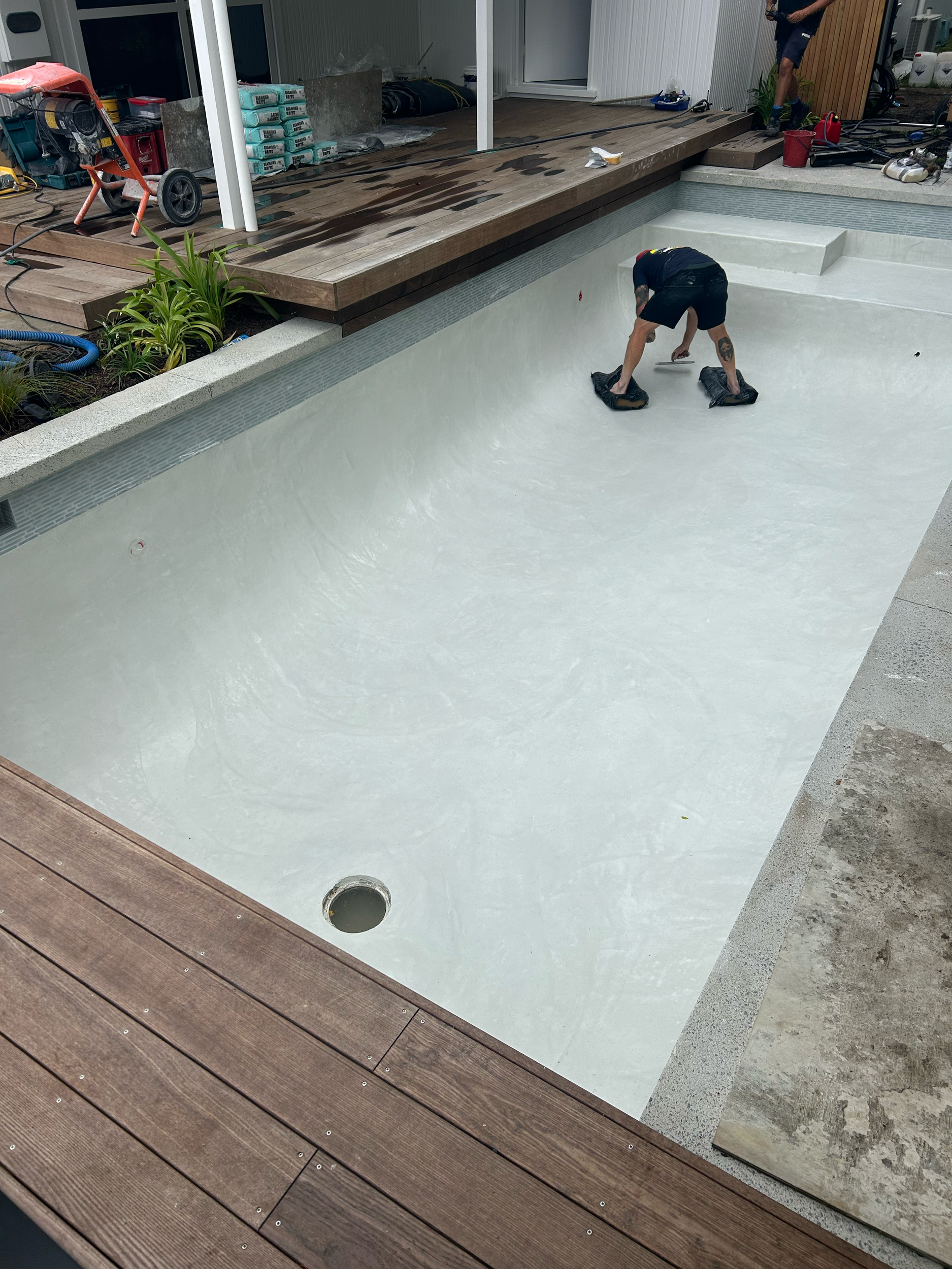 A person working on a swimming pool with a white interior, surrounded by wooden decking, tools, and construction supplies. Existing pool refurbishment Built by ESC outdoor living swimming pool builders. Auckland. skilled builders