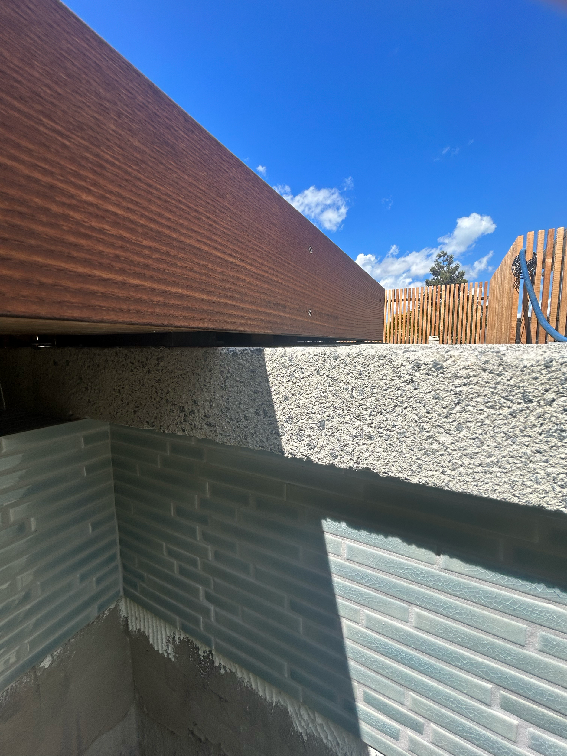 Close-up of a pool corner with a wooden railing, rough textured concrete block, and a greenish brick wall under a blue sky with some clouds. Existing pool refurbishment Built by ESC outdoor living swimming pool builders. Auckland. skilled builder