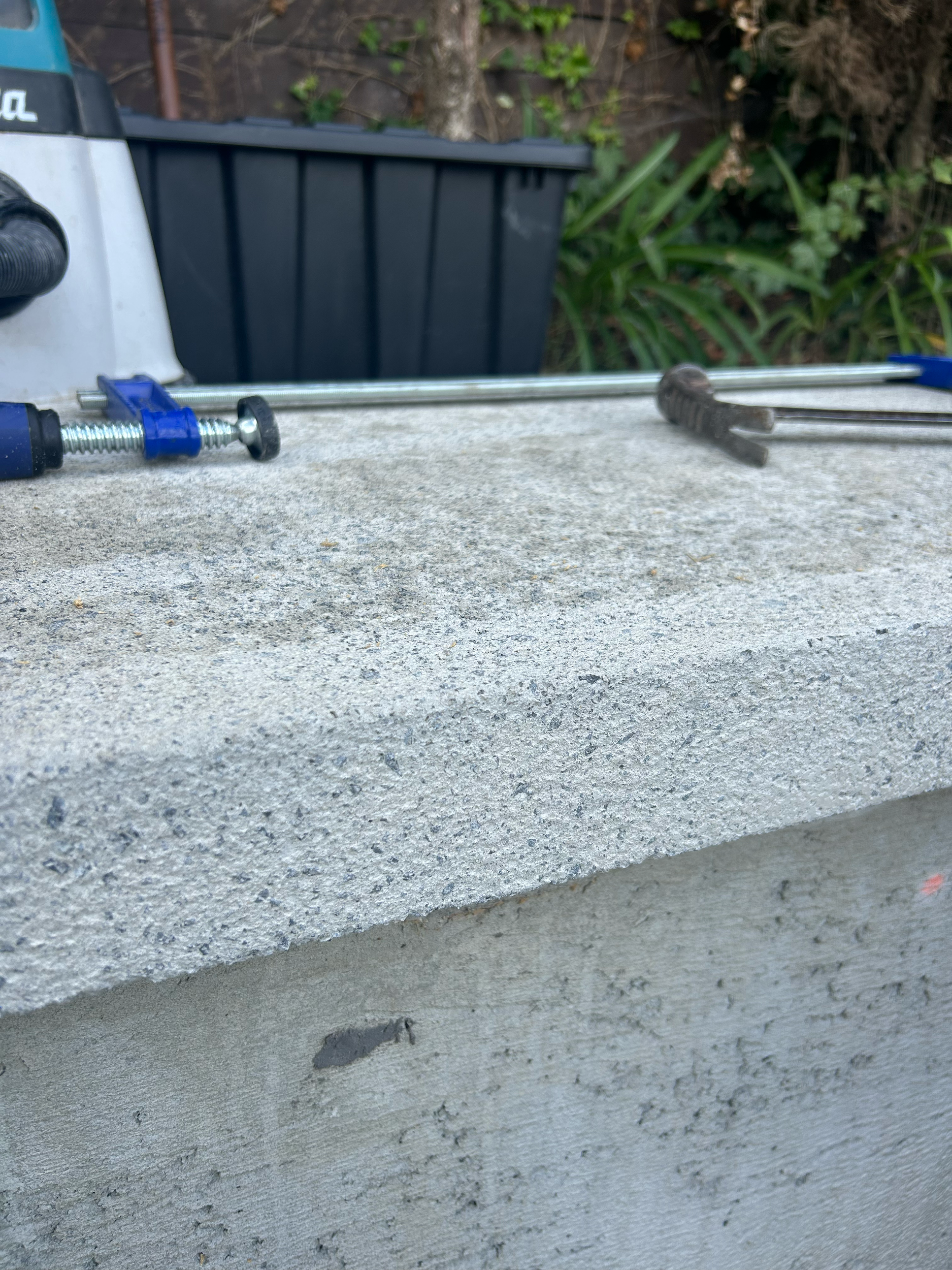 Tools on a concrete surface, including a clamp, a pry bar, and a long metal rod, with a black gardening container and green plants in the background. Existing pool refurbishment Built by ESC outdoor living swimming pool builders. Auckland.