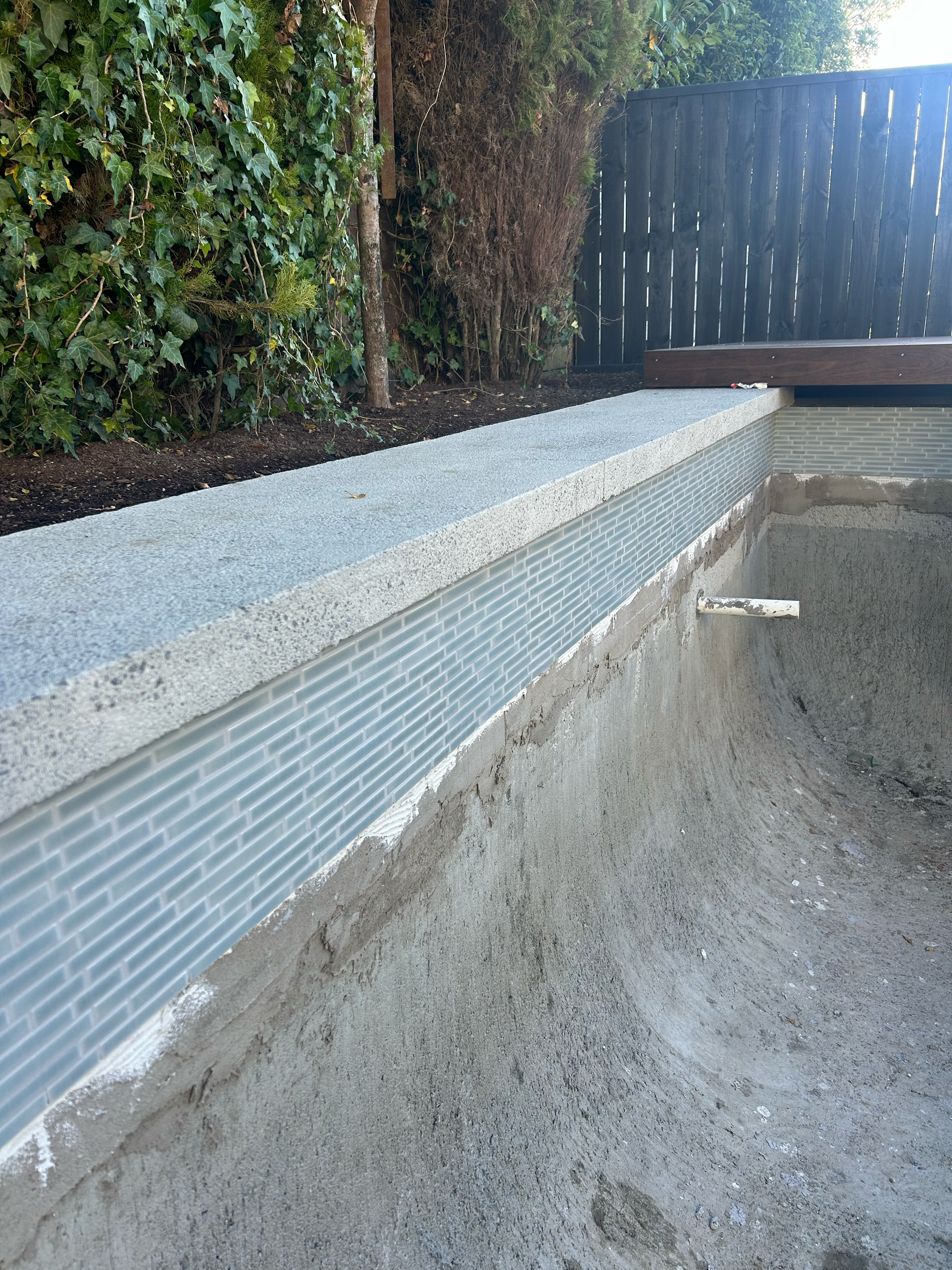 A concrete pool with a smooth, curved interior surface, surrounded by a gray gravel-like textured border and a flower bed with plants and trees behind a dark wooden privacy fence. Existing pool refurbishment Built by ESC outdoor living swimming pool