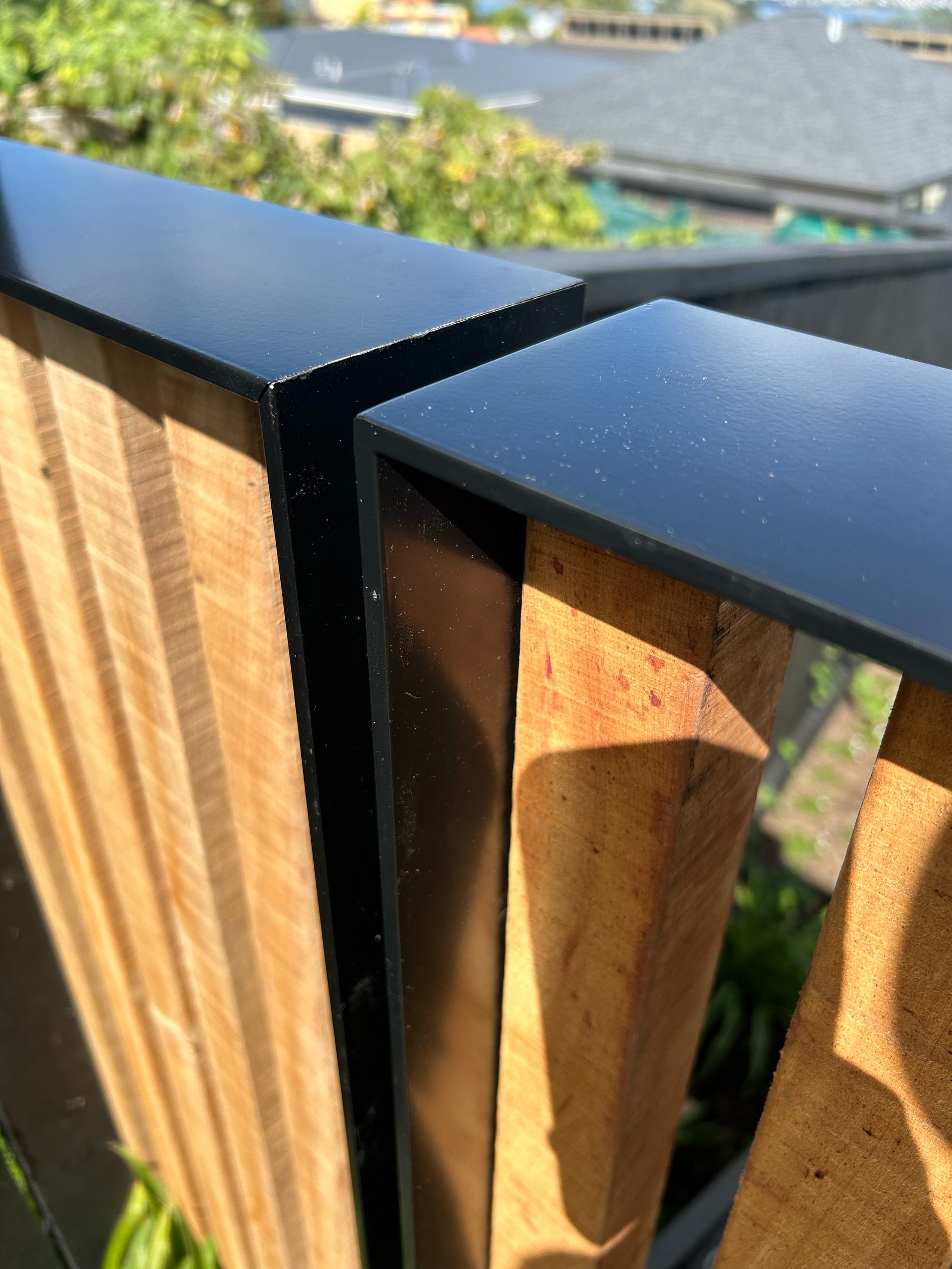Close-up of a wooden and metal railing on a balcony with a blurred view of rooftops and trees in the background. Existing pool refurbishment Built by ESC outdoor living swimming pool builders. Auckland. skilled builders