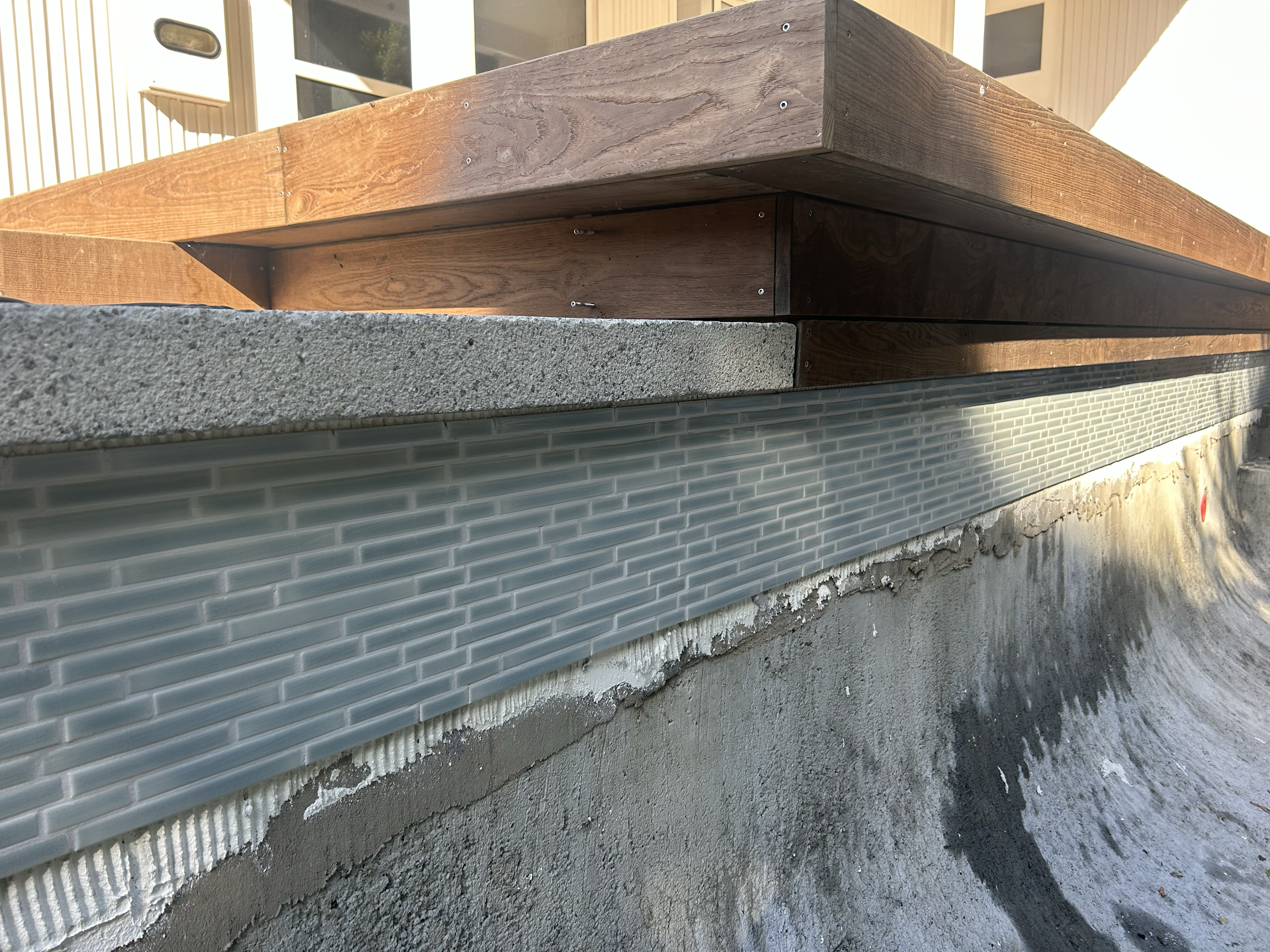 Close-up of a swimming pool, a textured concrete, a strip of small blue-green brick tiles, and a concrete surface, with a wooden deck above. Existing pool refurbishment Built by ESC outdoor living swimming pool builders. Auckland. skilled builders