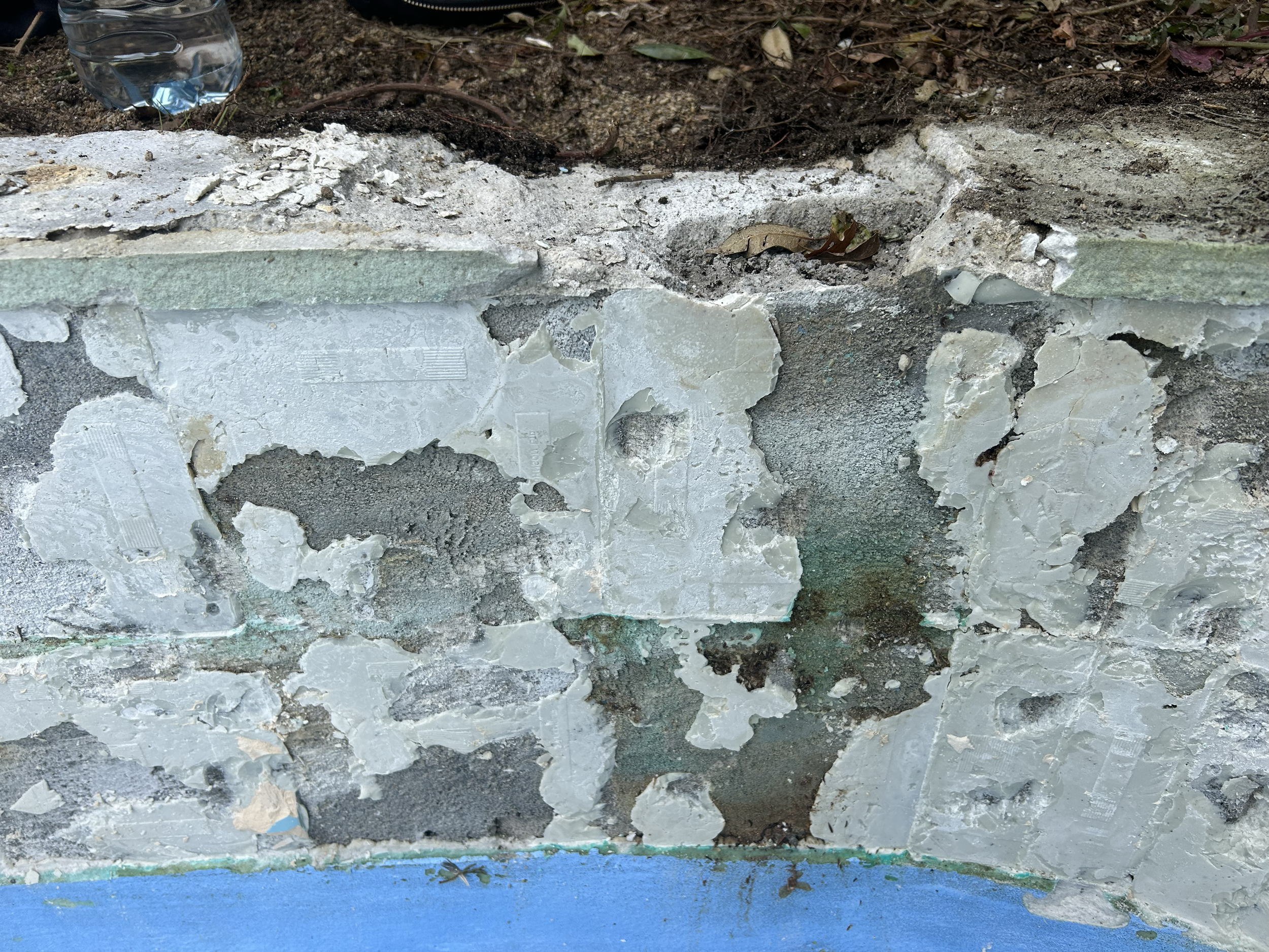 Close-up of a damaged concrete wall with peeling paint and exposed rough surface, with soil and a plastic bottle in the background. Existing pool refurbishment Built by ESC outdoor living swimming pool builders. Auckland.
