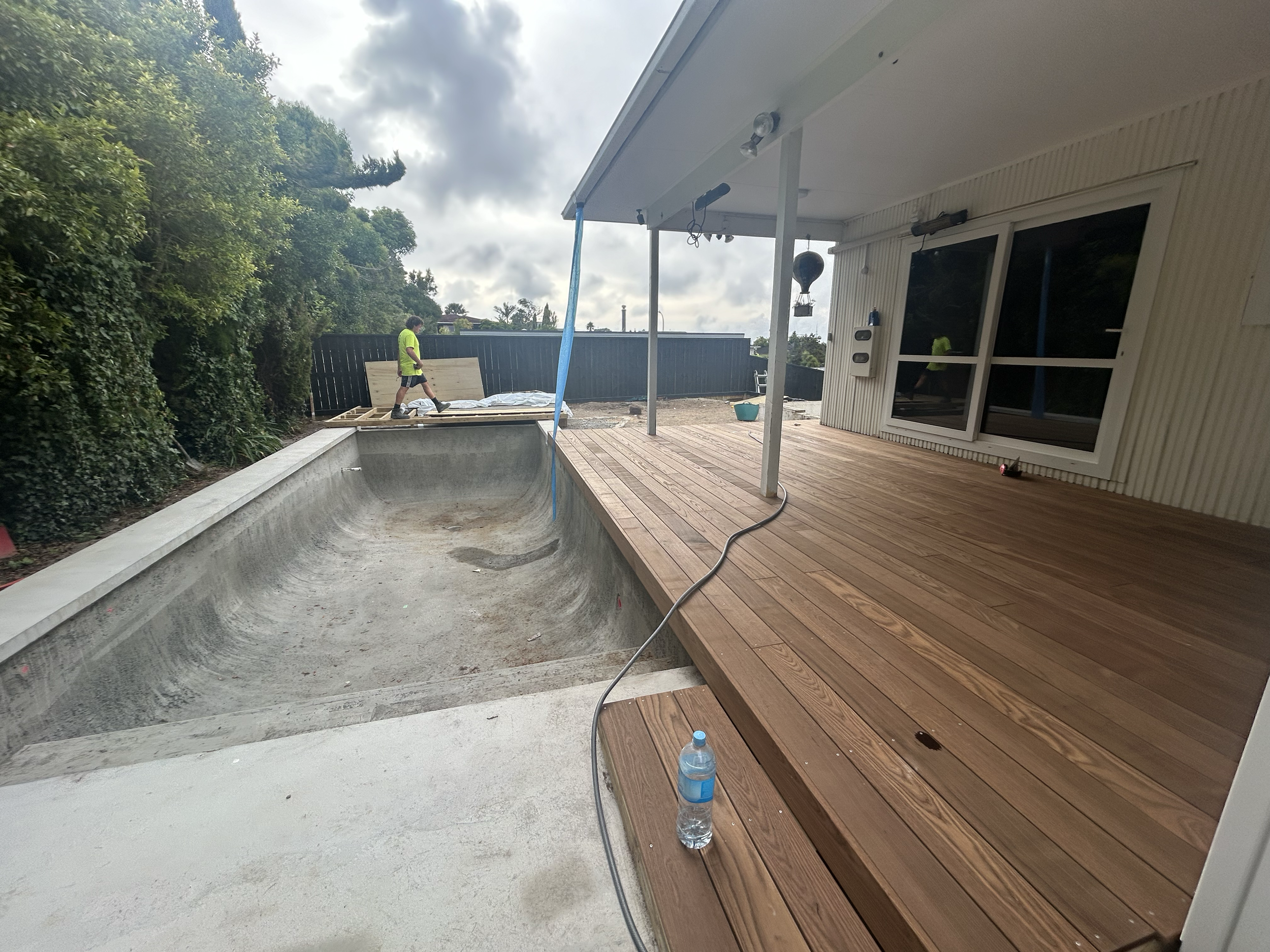 Undergoing construction on backyard pool with adjacent wooden deck, with two workers and construction materials present. Existing pool refurbishment Built by ESC outdoor living swimming pool builders. Auckland. skilled builders