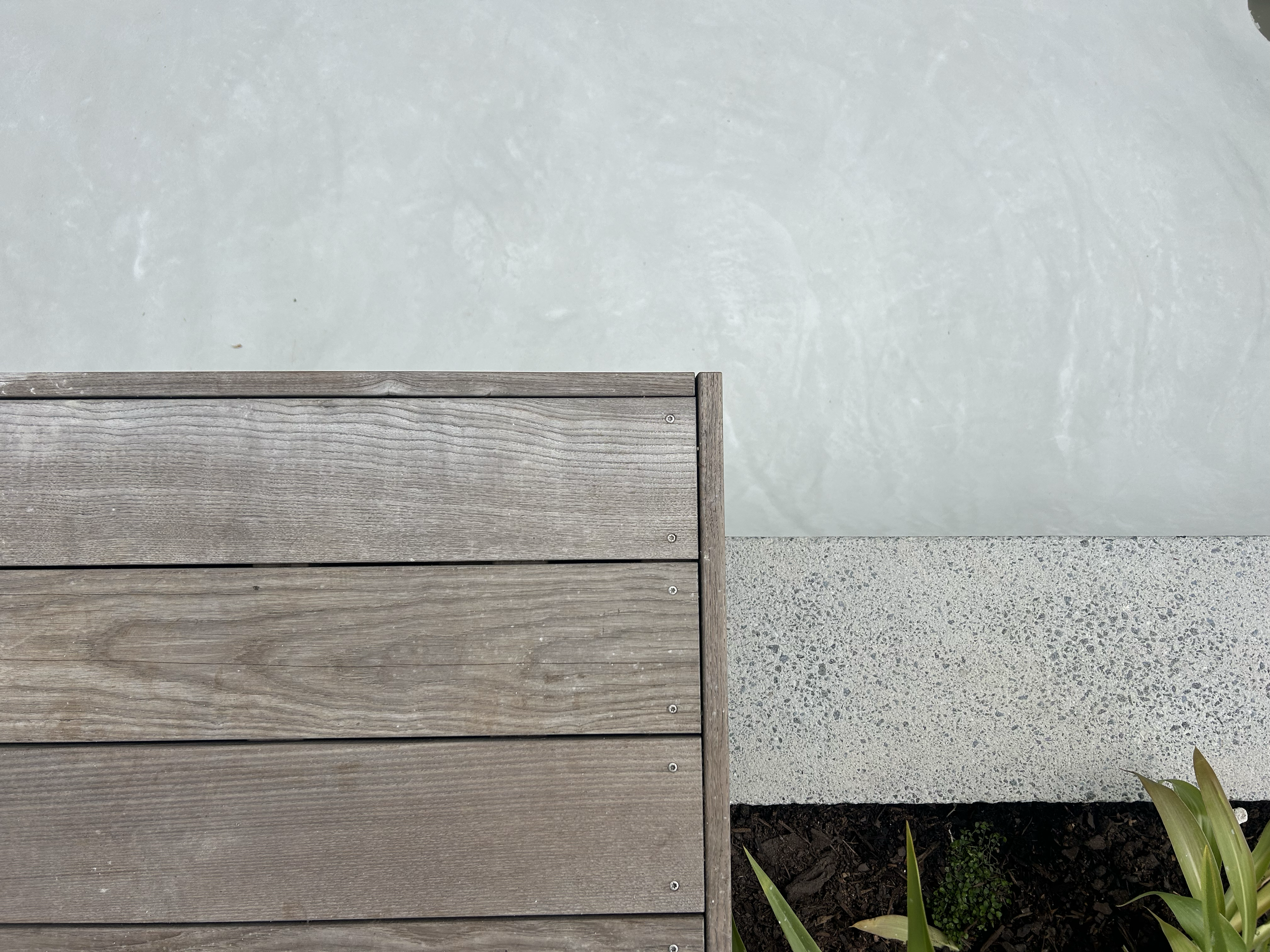 Close-up of a wooden bench on a concrete surface with a plant in the bottom right corner. Existing pool refurbishment Built by ESC outdoor living swimming pool builders. Auckland. skilled builders
