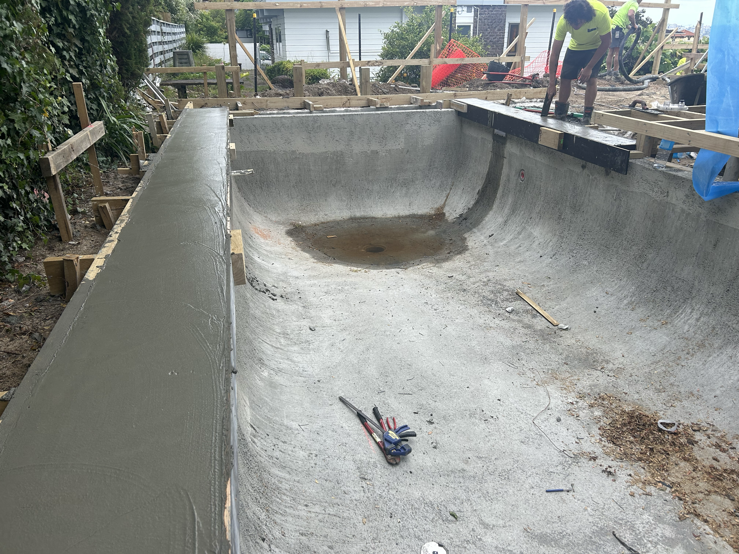 Under construction backyard swimming pool with new concrete surface, with workers working around the edge. Existing pool refurbishment Built by ESC outdoor living swimming pool builders. Auckland.