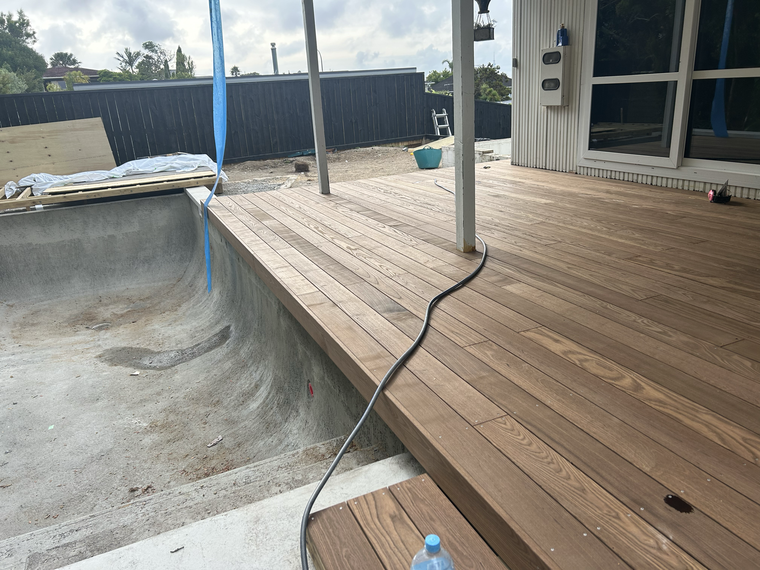 Construction of a wooden deck next to an empty swimming pool. The deck has a pole with a canopy, construction tools, and materials visible around the area. Existing pool refurbishment Built by ESC outdoor living swimming pool builders. Auckland.