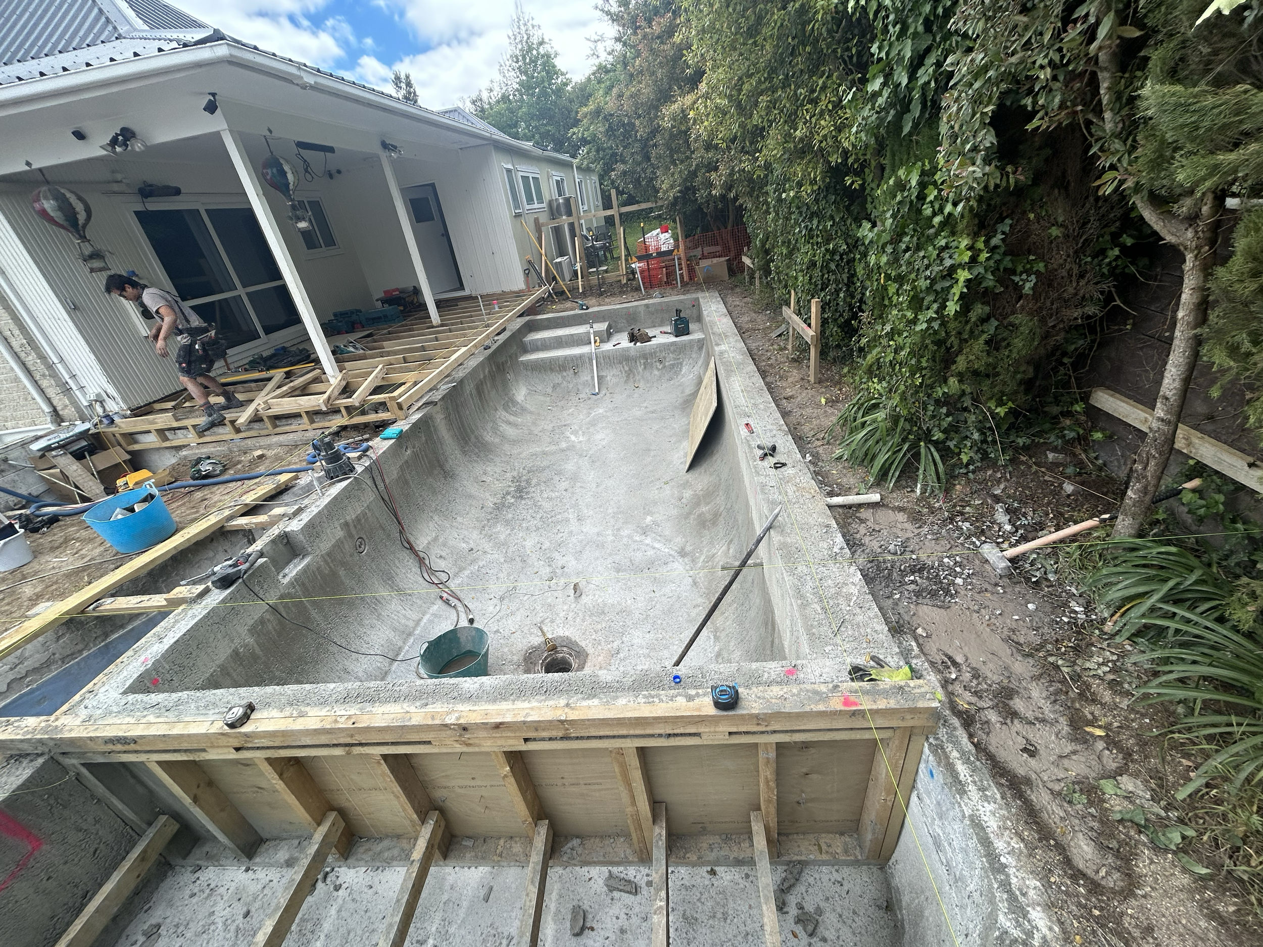 Under construction backyard swimming pool with wooden framing, construction tools, and workers working on the deck area adjacent to the pool. Surrounding trees and bushes in the background. Existing pool refurbishment Built by ESC outdoor living swim