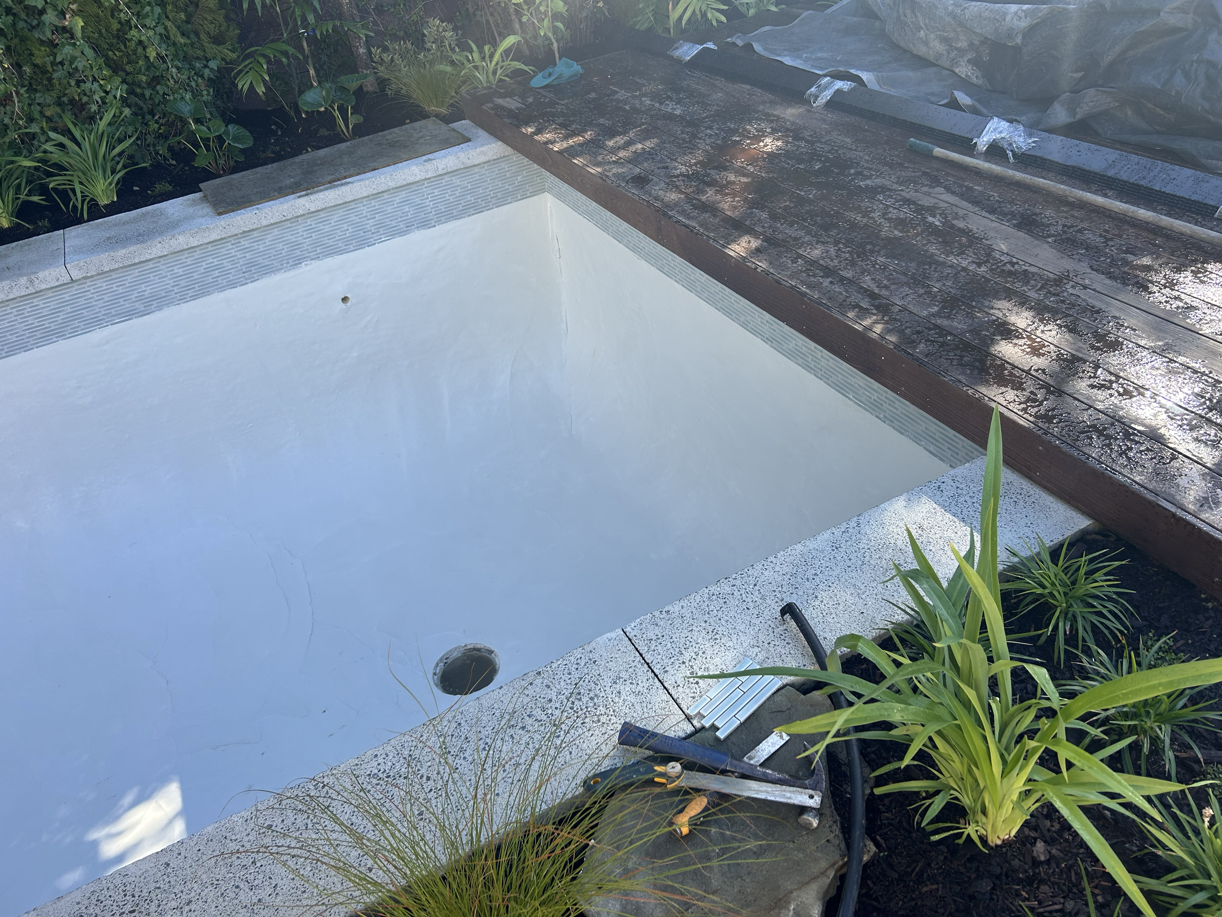 Empty swimming pool with a drilled drain and surrounded by plants, with construction tools placed nearby. Existing pool refurbishment Built by ESC outdoor living swimming pool builders. Auckland. skilled builders
