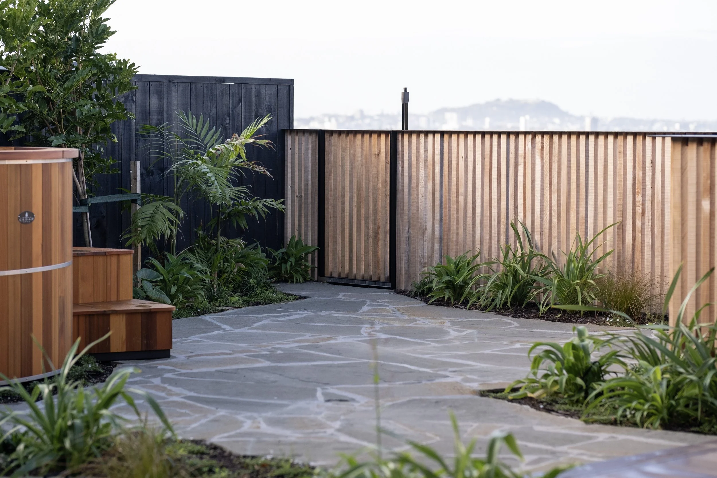 A rooftop garden with stone pathway, green plants and wooden fencing, with cityscape in the distance. Existing pool refurbishment Built by ESC outdoor living swimming pool builders. Auckland. skilled builders
