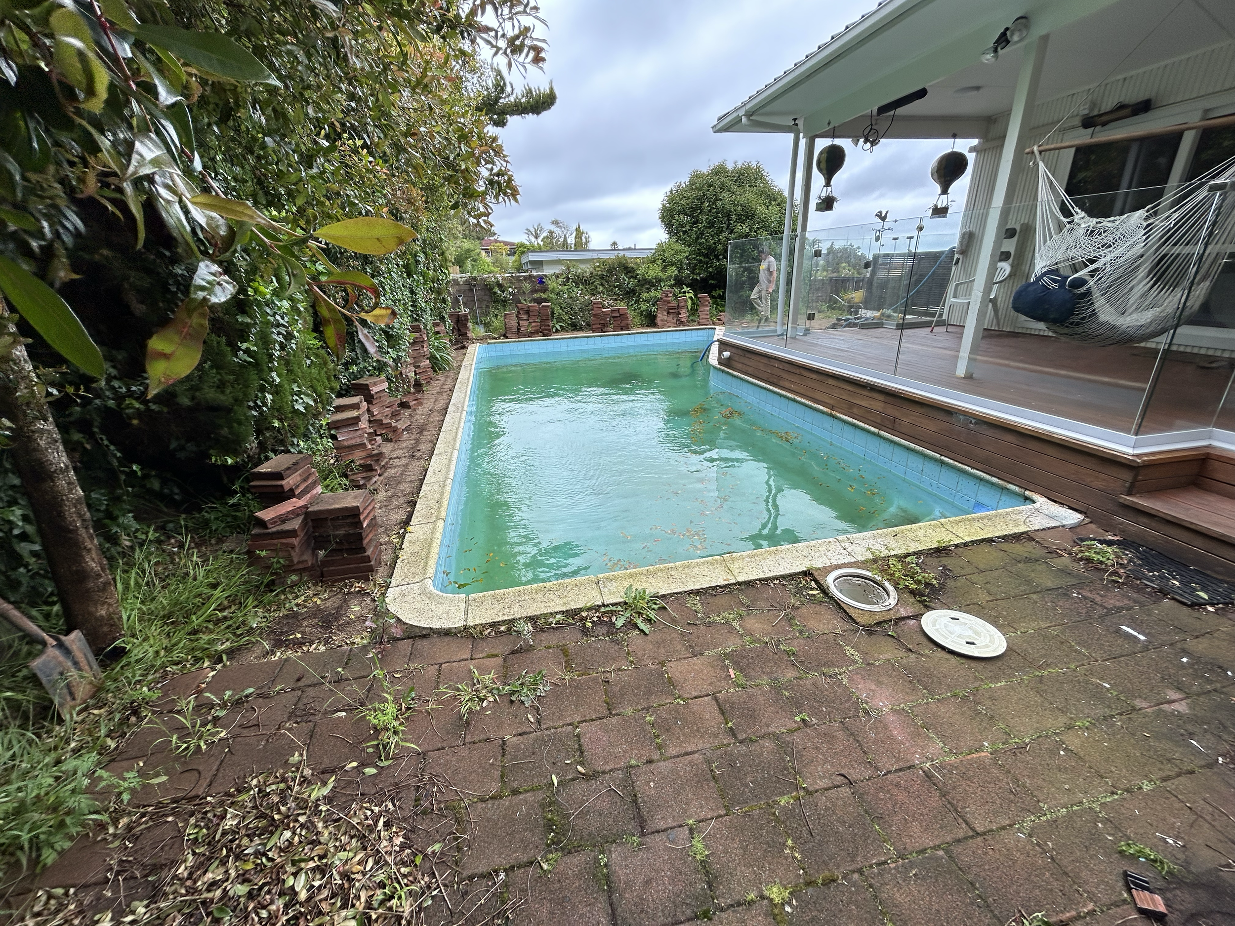 A backyard featuring a small, rectangular swimming pool with dirty water and algae. Surrounding the pool are brick pavers, some overgrown with weeds. Existing pool refurbishment Built by ESC outdoor living swimming pool builders. Auckland.