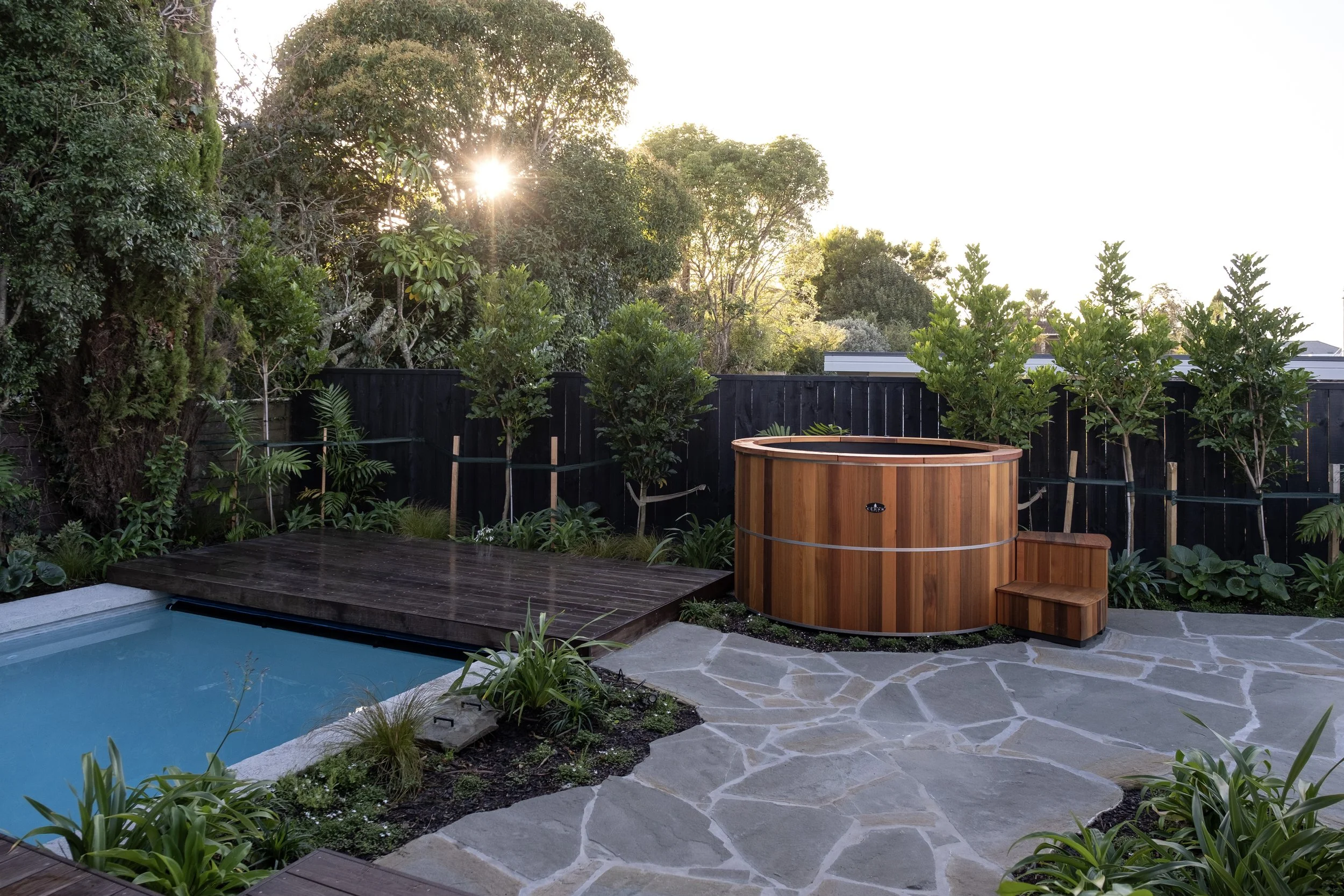 Backyard with a hot tub, small pool, wooden deck, stone patio, and lush trees and plants in the background. Existing pool refurbishment Built by ESC outdoor living swimming pool builders. Auckland. skilled builders