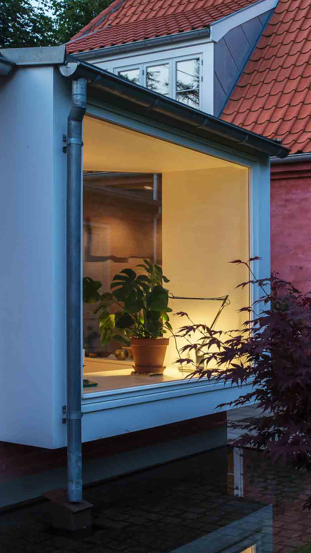 Et vindue med indsigt prydet af en potteplante, belyst med varmt lys, på en bygning med rød tegltag og blå facade, omgivet af træer.