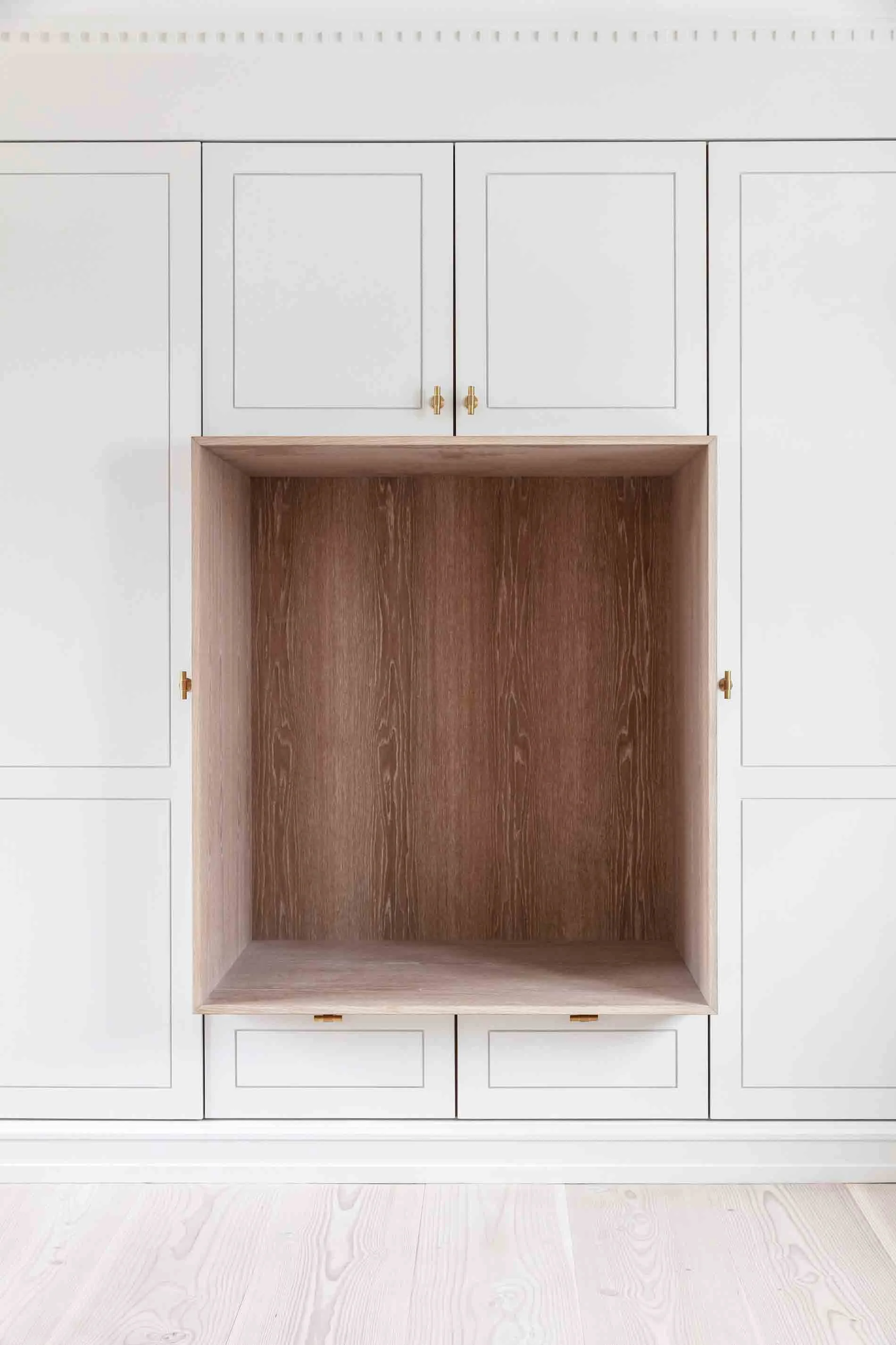 Et tomt, indbygget vaskeskab eller hyldesystem med et naturligt træinteriør, omgivet af hvide skabe med guld håndtag.