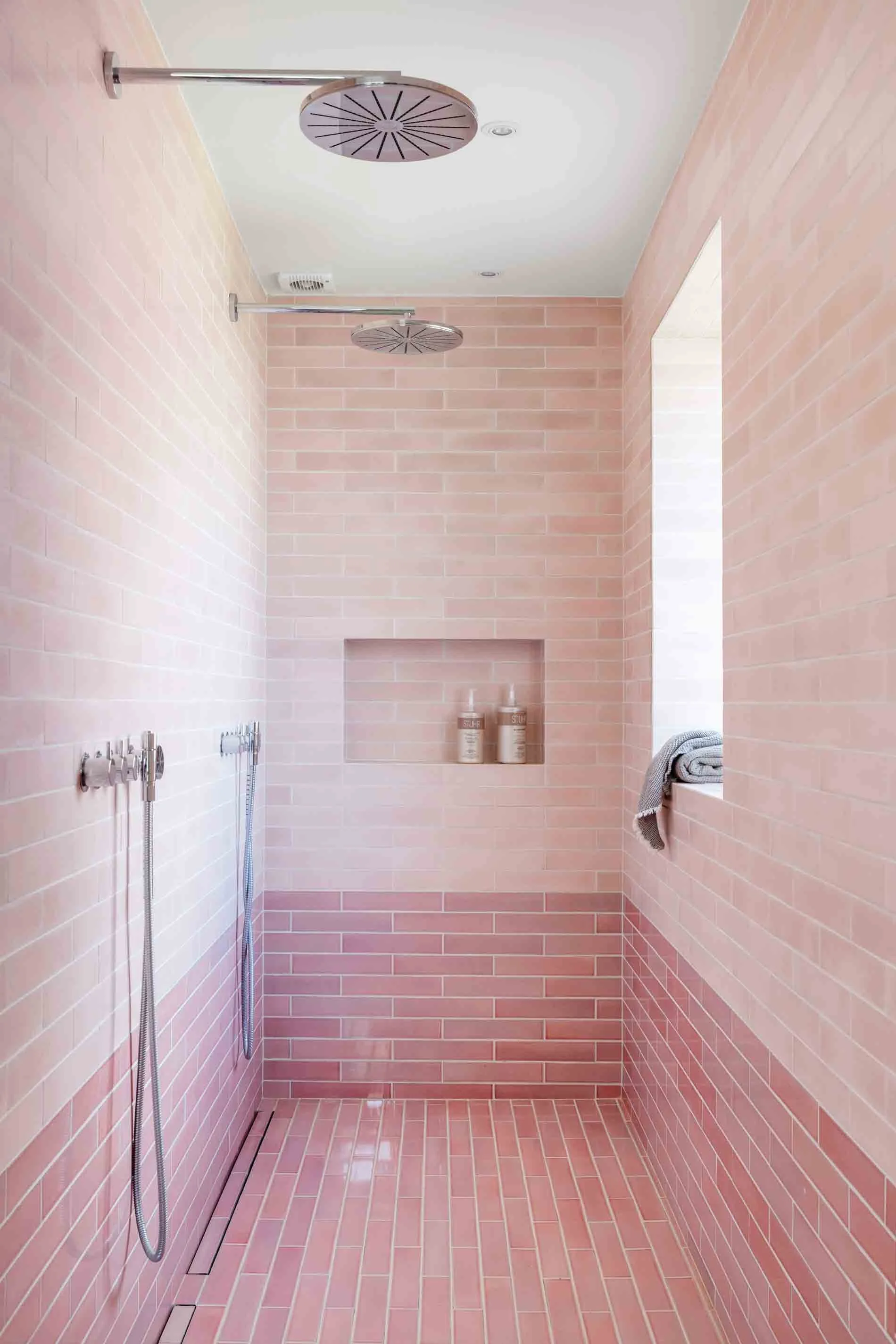 Moderne brusebad med rosa fliser, to regnbruser på loftet, og en niche med shampoo-Flasker. Der er en vindue med et håndklæde liggende over vindueskarmen.