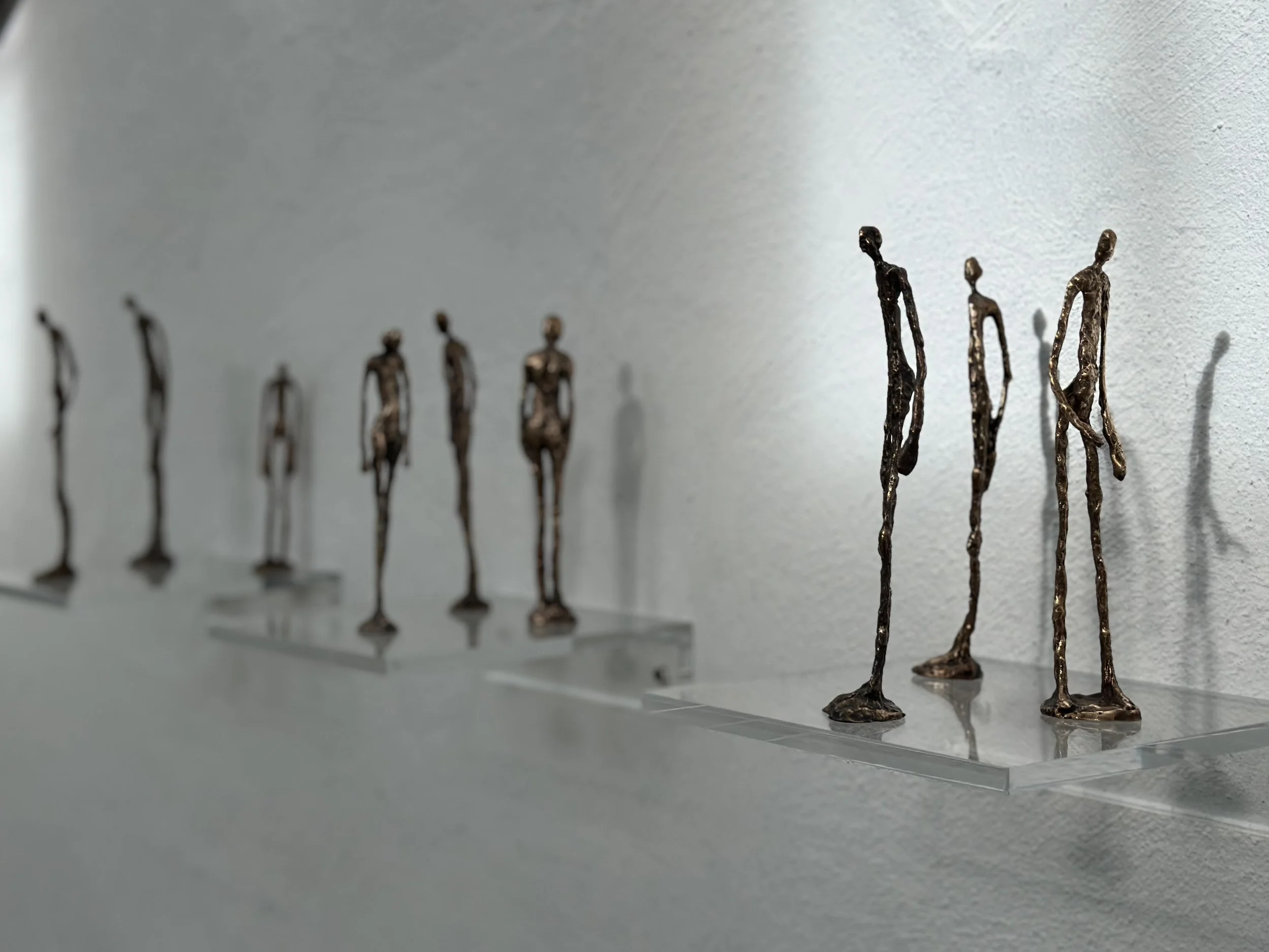 A row of abstract metal sculptures of elongated human figures lined up on a shelf against a plain wall.