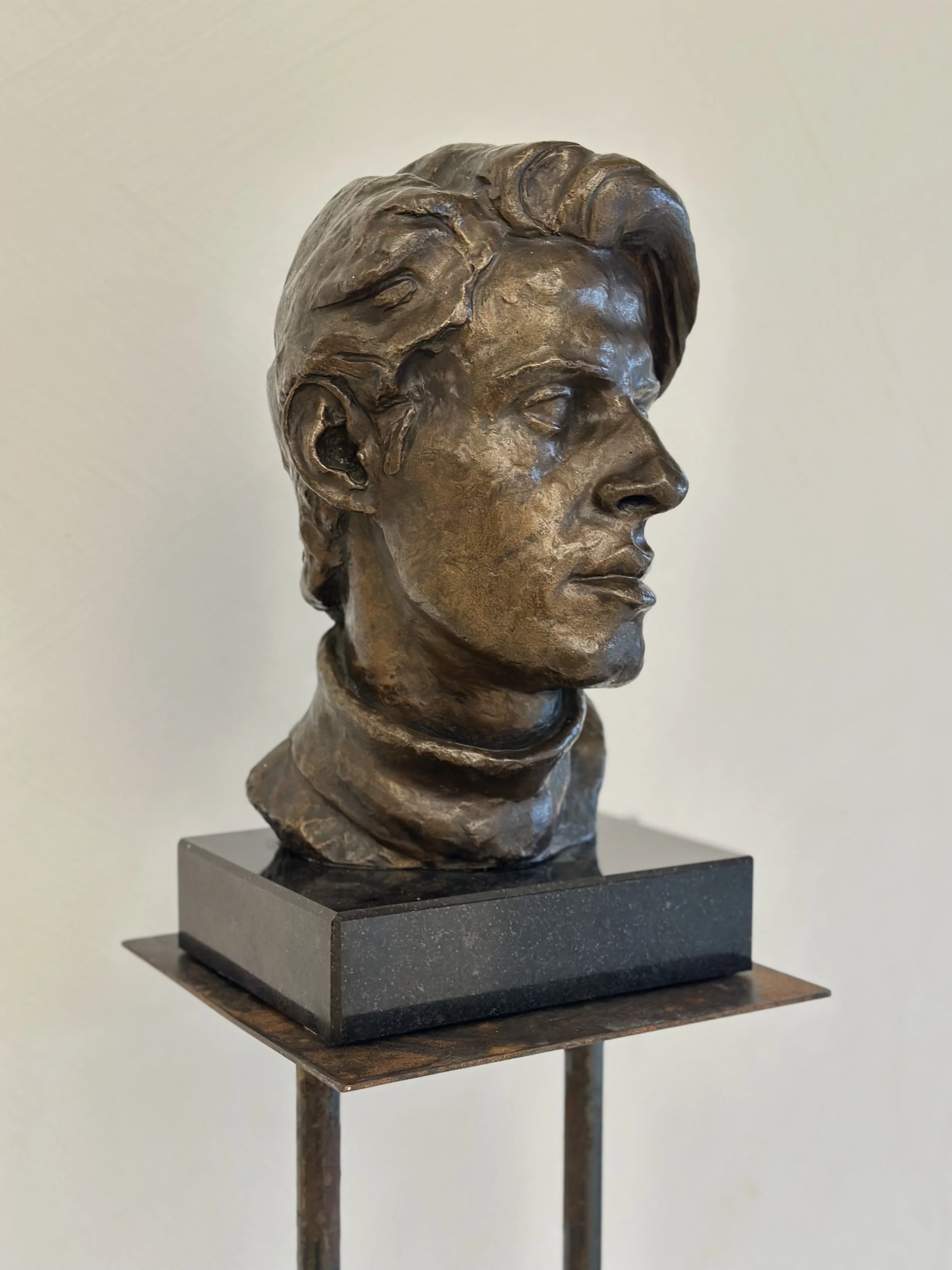 Bronze bust sculpture of a person's head facing right, mounted on a black rectangular base and a stand.