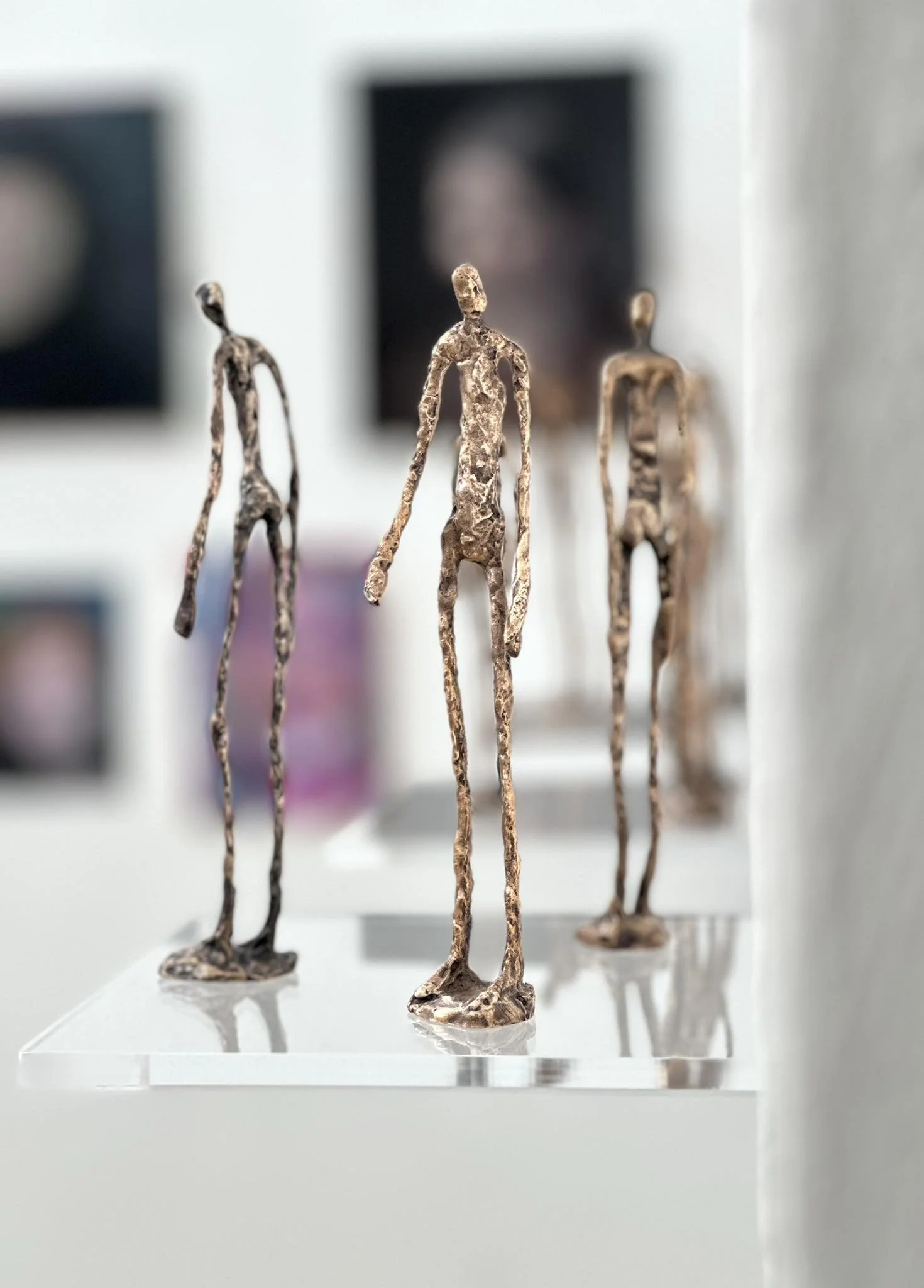 Three abstract metallic sculptures of elongated humanoid figures on a clear display stand in an art gallery.