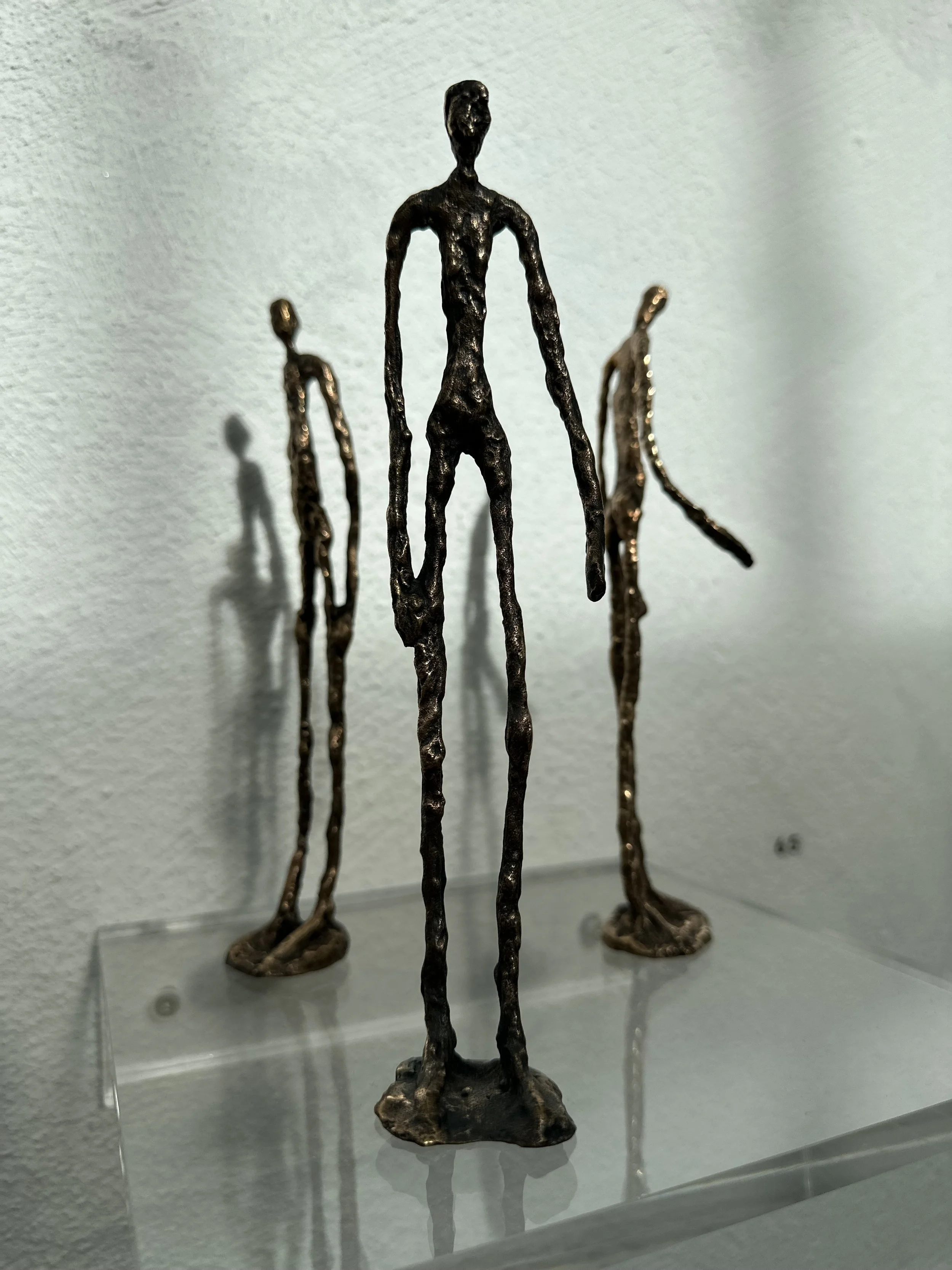 Three tall, abstract bronze sculptures of human figures with elongated limbs and minimal facial features, displayed on a clear platform against a white wall.