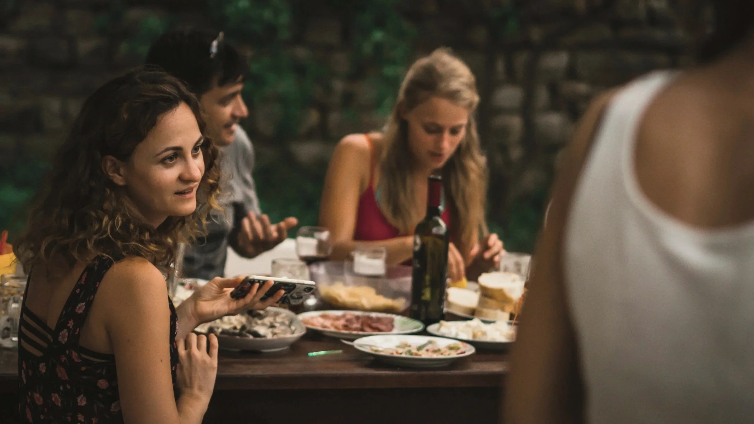 Group of people sitting around a dinner table outdoors, with food and a bottle of wine, engaging in conversation.