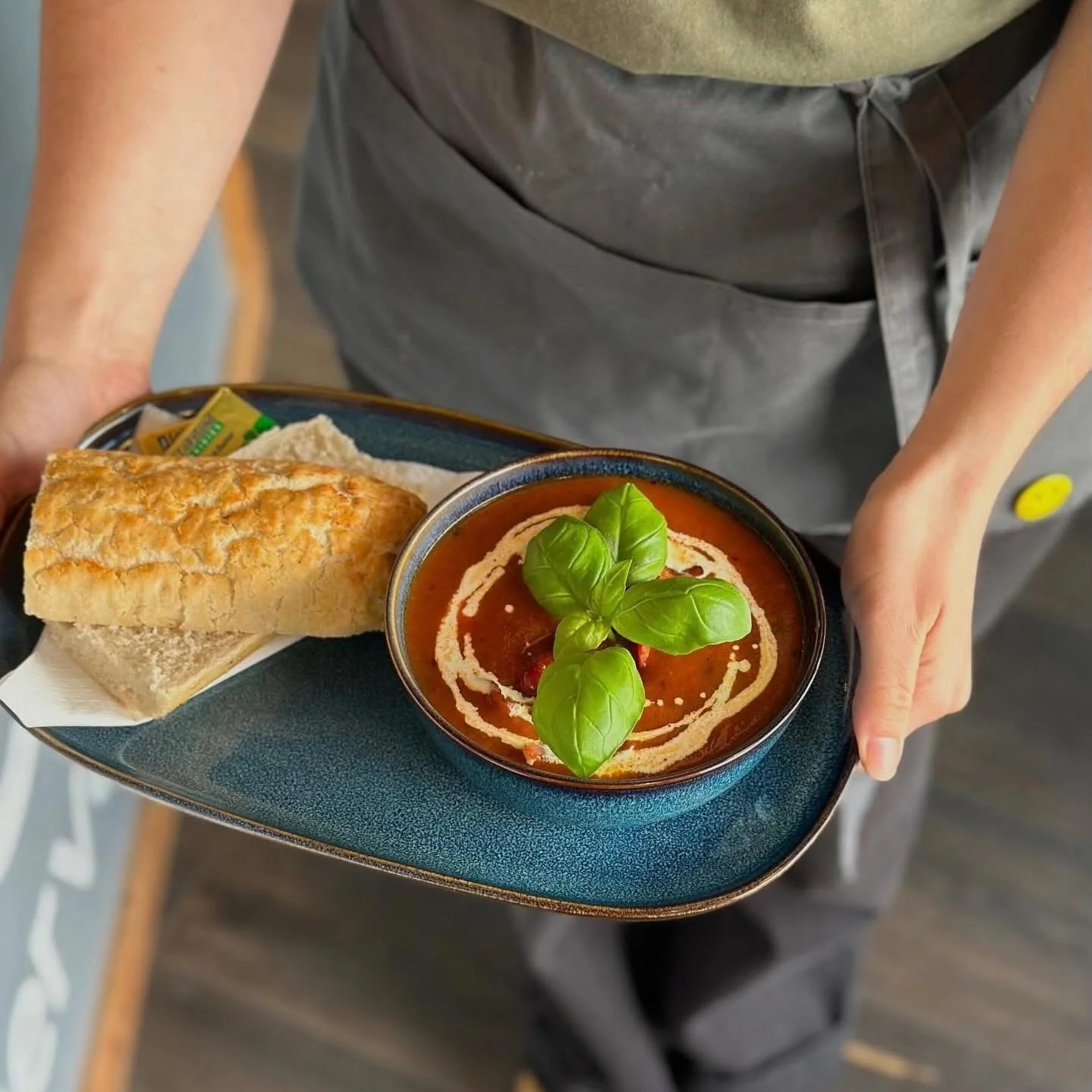 A top down view of a barista delivering a tasty tray of tomato soup, with fresh basil leaves and swirls of cream, next to some freshly baked crispy bread and butter