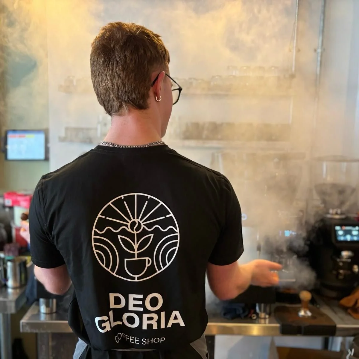 A barista making coffee with his back to the camera. His t-shirt shows the Deo Gloria logo