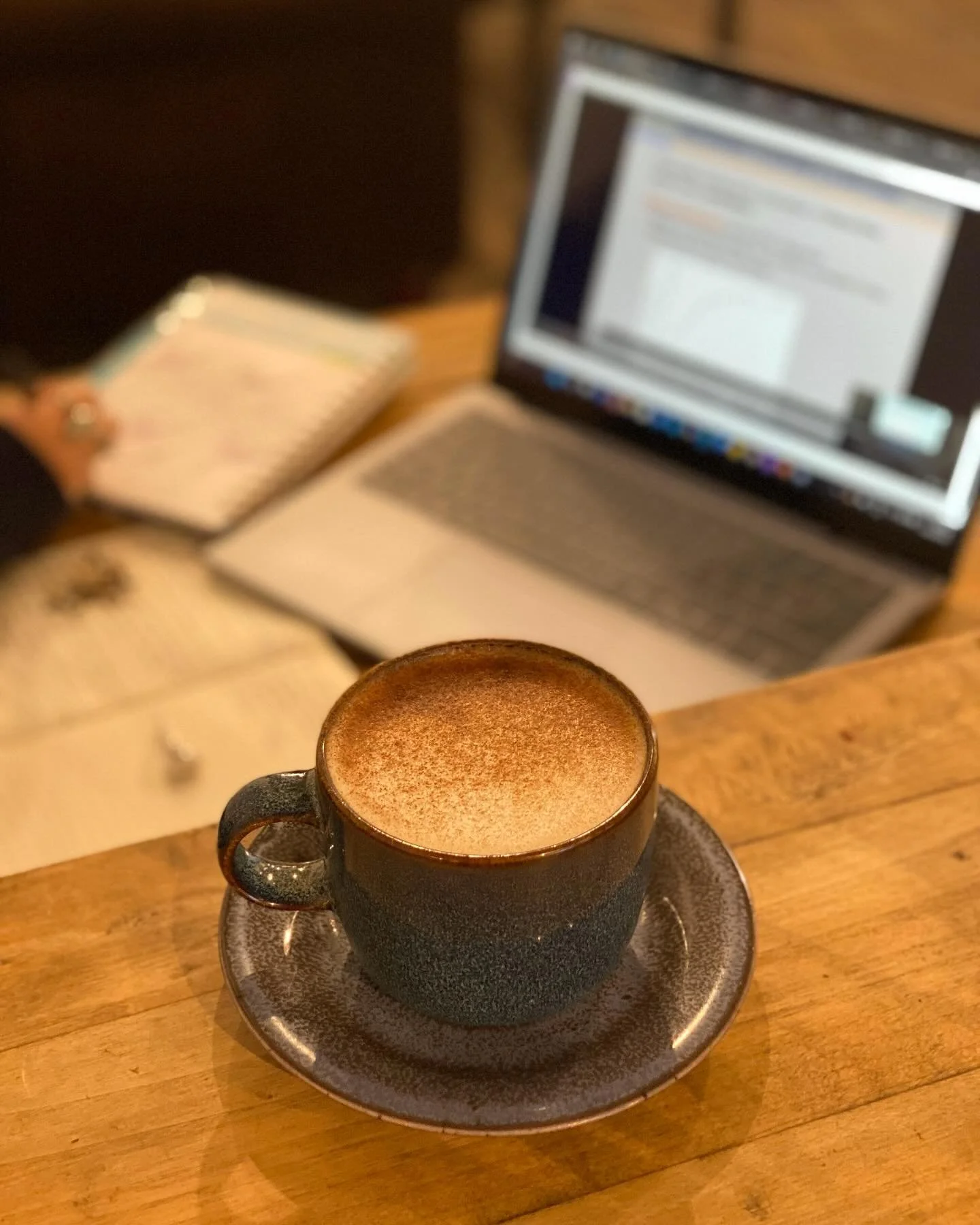 A freshly made coffee stands in the foreground with an open laptop and a persons hand and notebook are blurry in the background