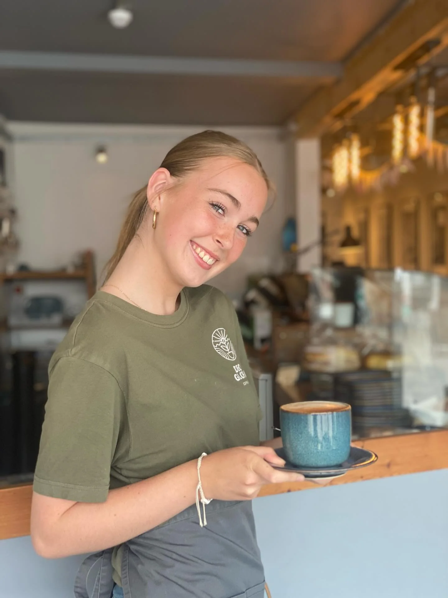 A barista on her way to deliver another delicious Deo Gloria latte smiles sweetly at the camera, her head tilted to her left