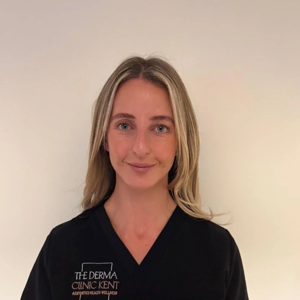 A woman with blonde hair wearing a black medical uniform with the words 'The Derma Clinic Kent' embroidered on it, standing against a plain beige background.