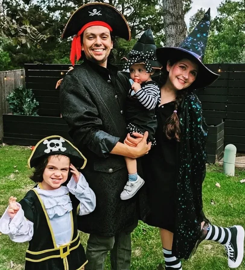 A family dressed in Halloween costumes outdoors, including a pirate, a witch, a little girl with a pirate hat, and a baby dressed as a witch, all smiling.