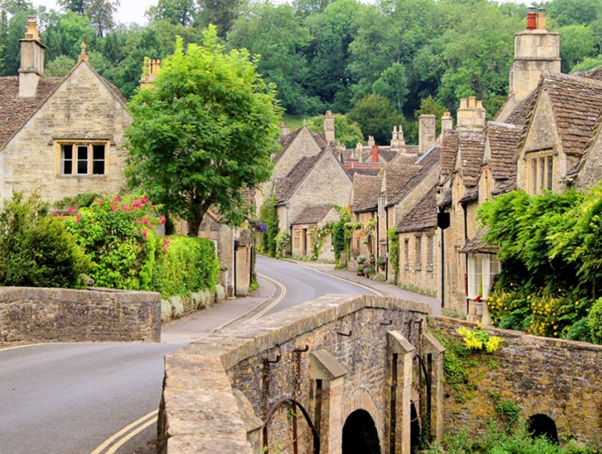 Charming village street with stone houses, lush greenery, and a small stone bridge.