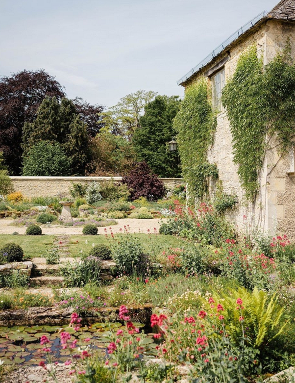 A lush garden with various flowering plants, bushes, and trees next to an old stone house covered in climbing ivy, with a stone pathway and a pond with water lilies.