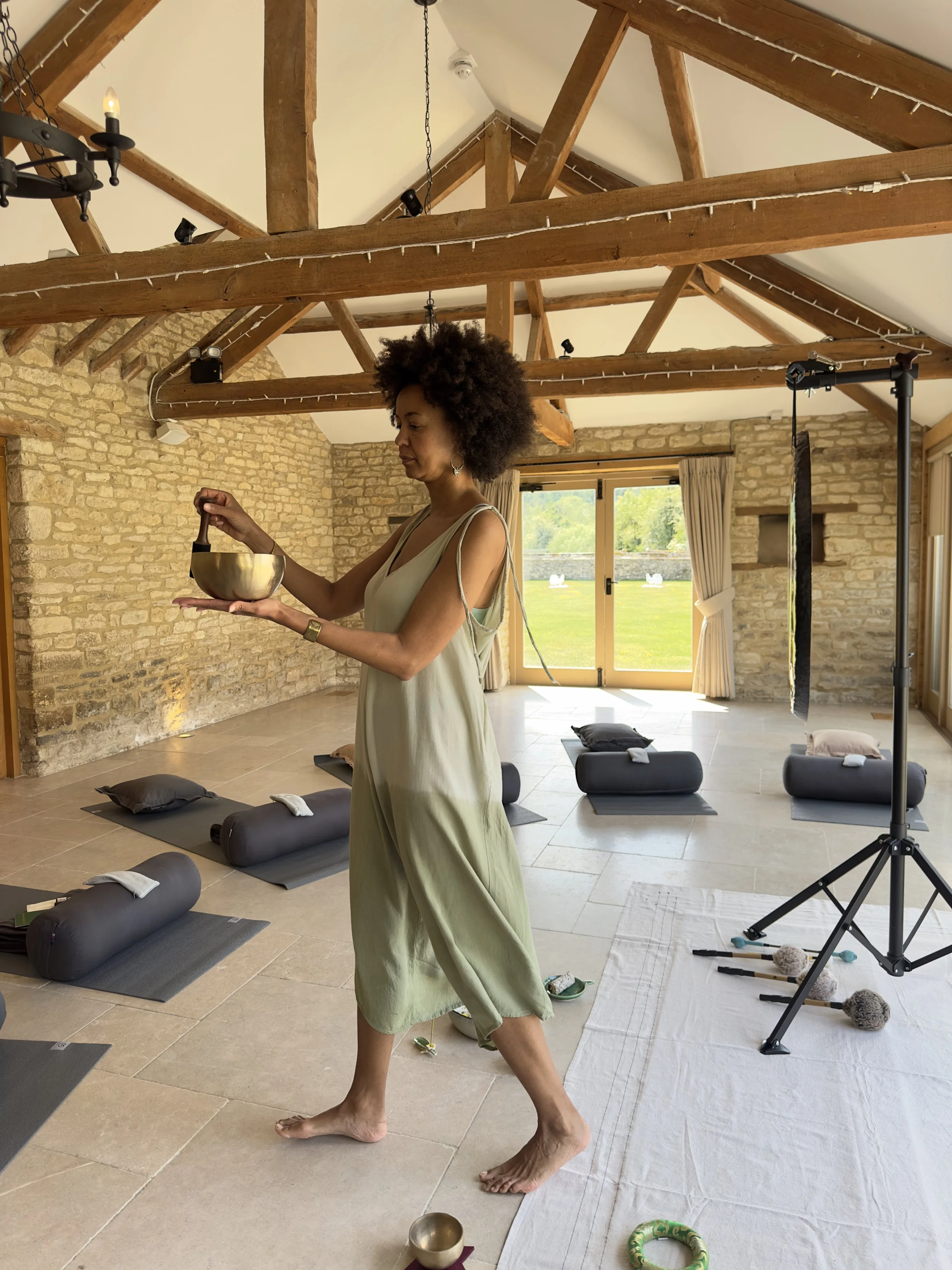 Woman preparing singing bowls in a yoga or meditation studio with mats, bolsters, and supplies, in a room with exposed wooden beams and a stone wall.