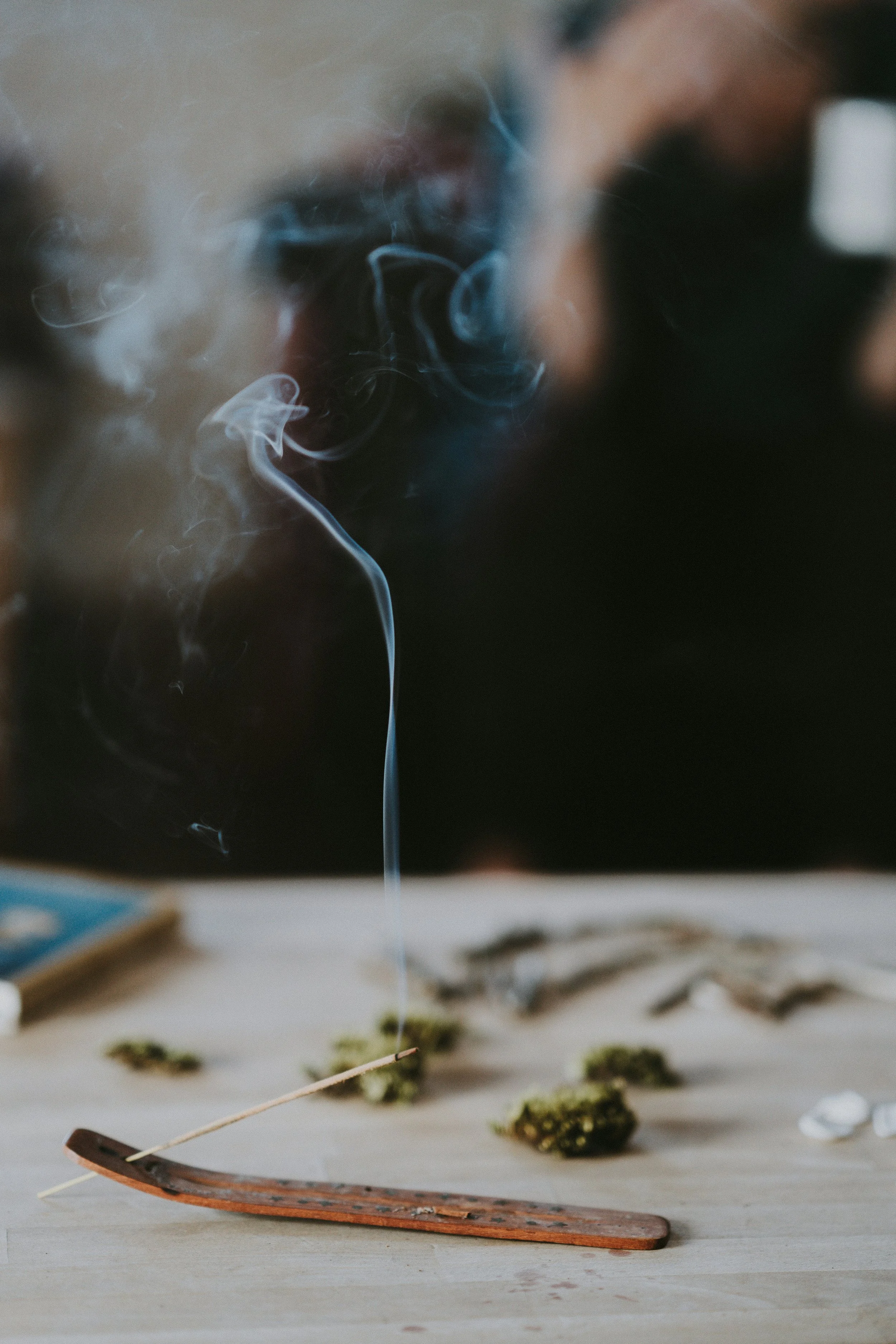 Spilled cannabis buds, a burning wooden stick, and smoking incense on a table with a person's face blurred in the background.