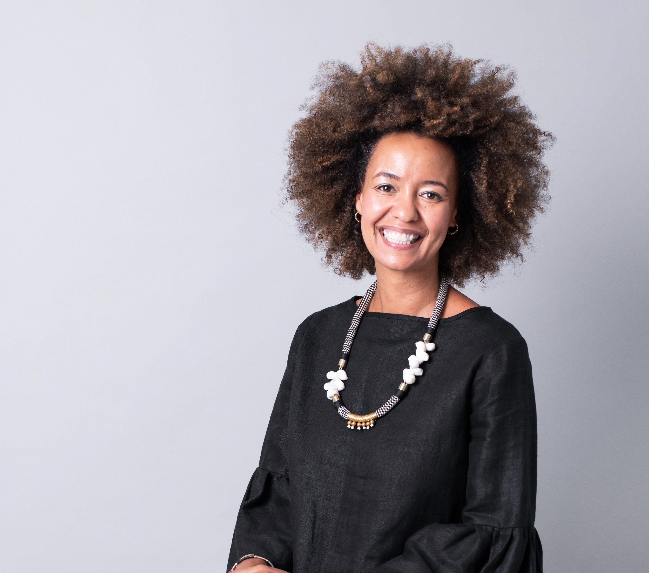 Smiling woman with curly brown hair wearing a black top and a statement necklace, standing against a plain light gray background.
