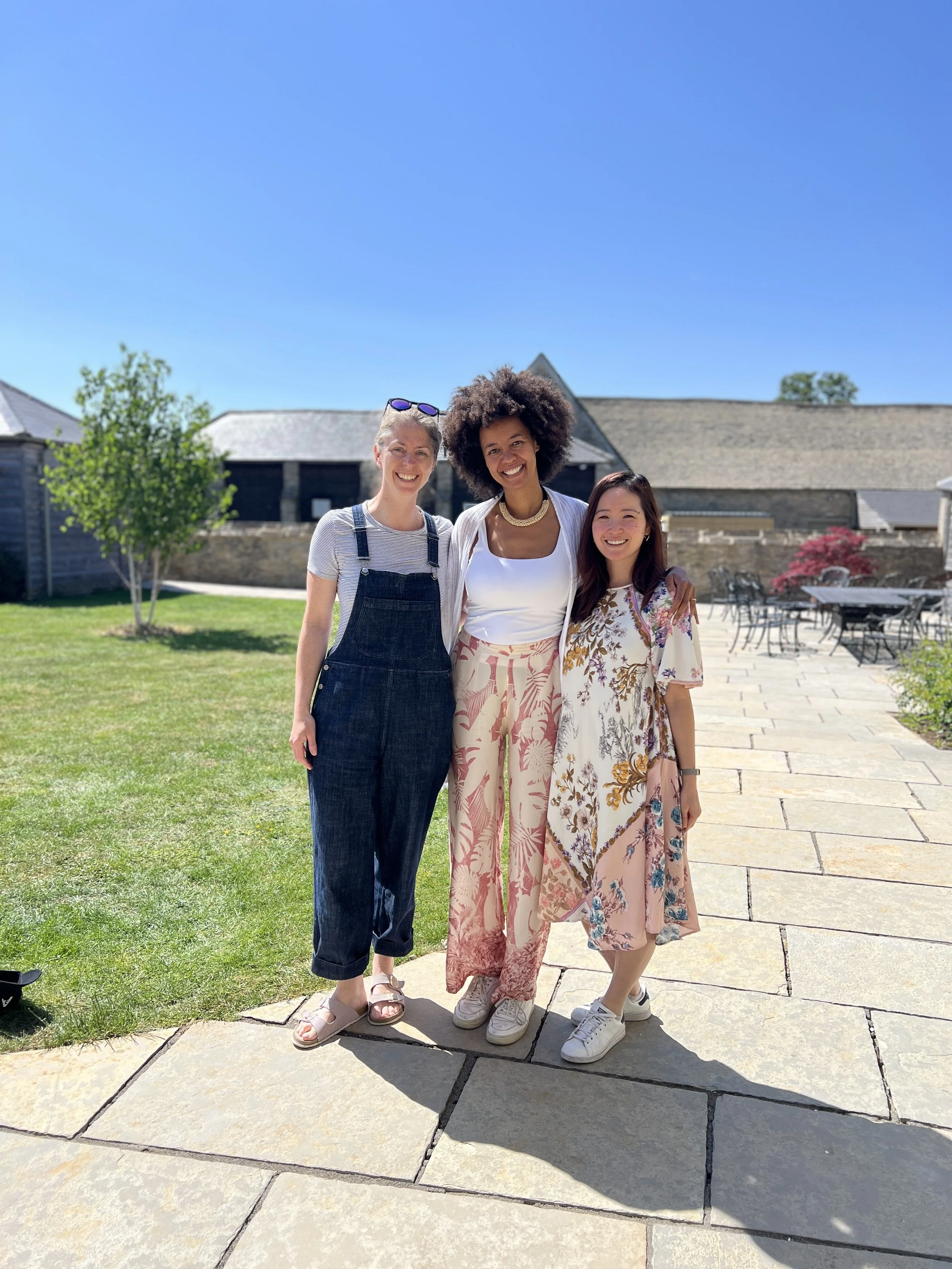 Three women standing outdoors on a sunny day, smiling, with a grassy area, trees, and buildings in the background. hosting a retreat in the cotswolds