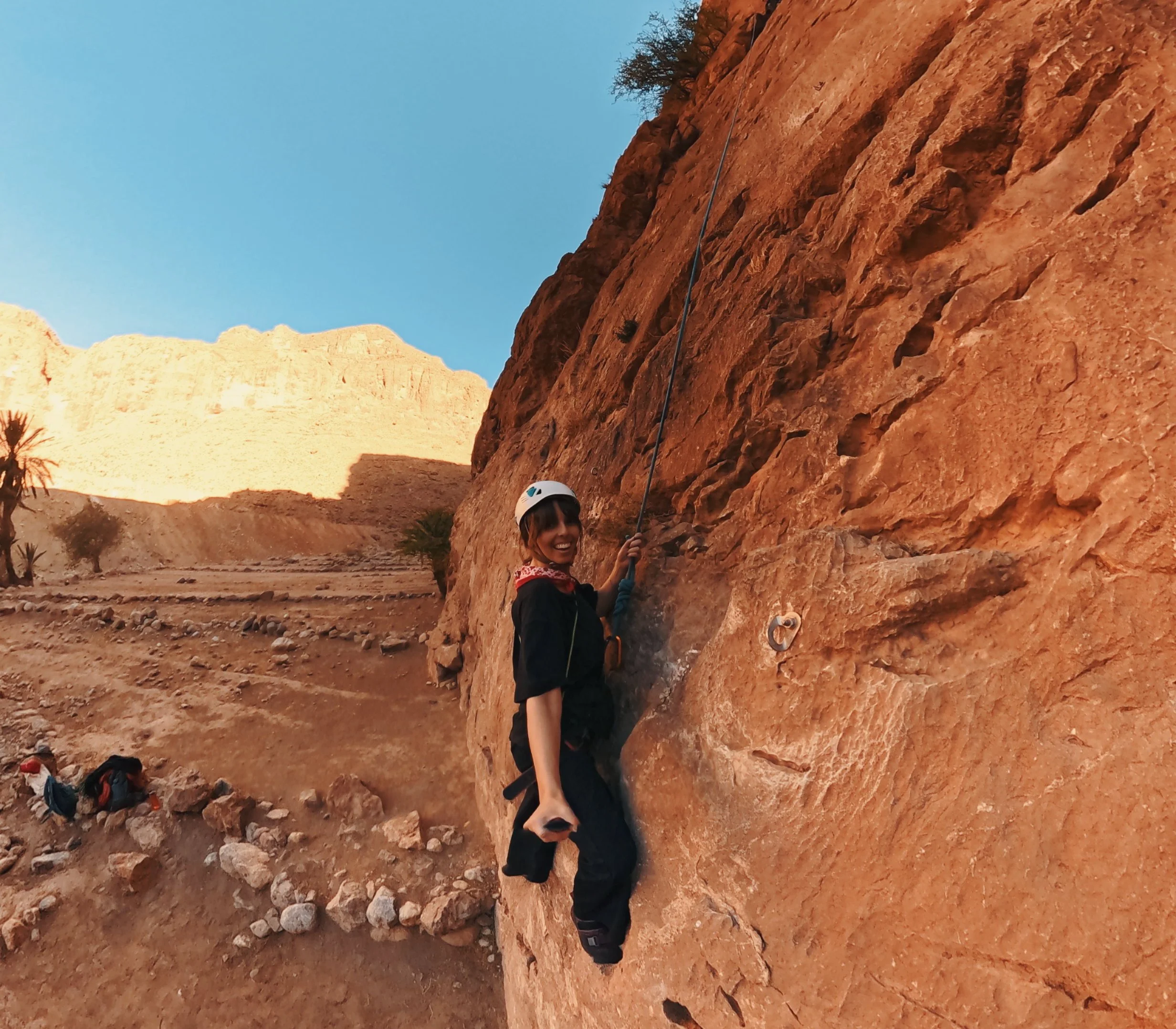 A woman wearing a helmet and climbing harness is rock climbing on a red sandstone cliff in a desert landscape with clear blue sky.