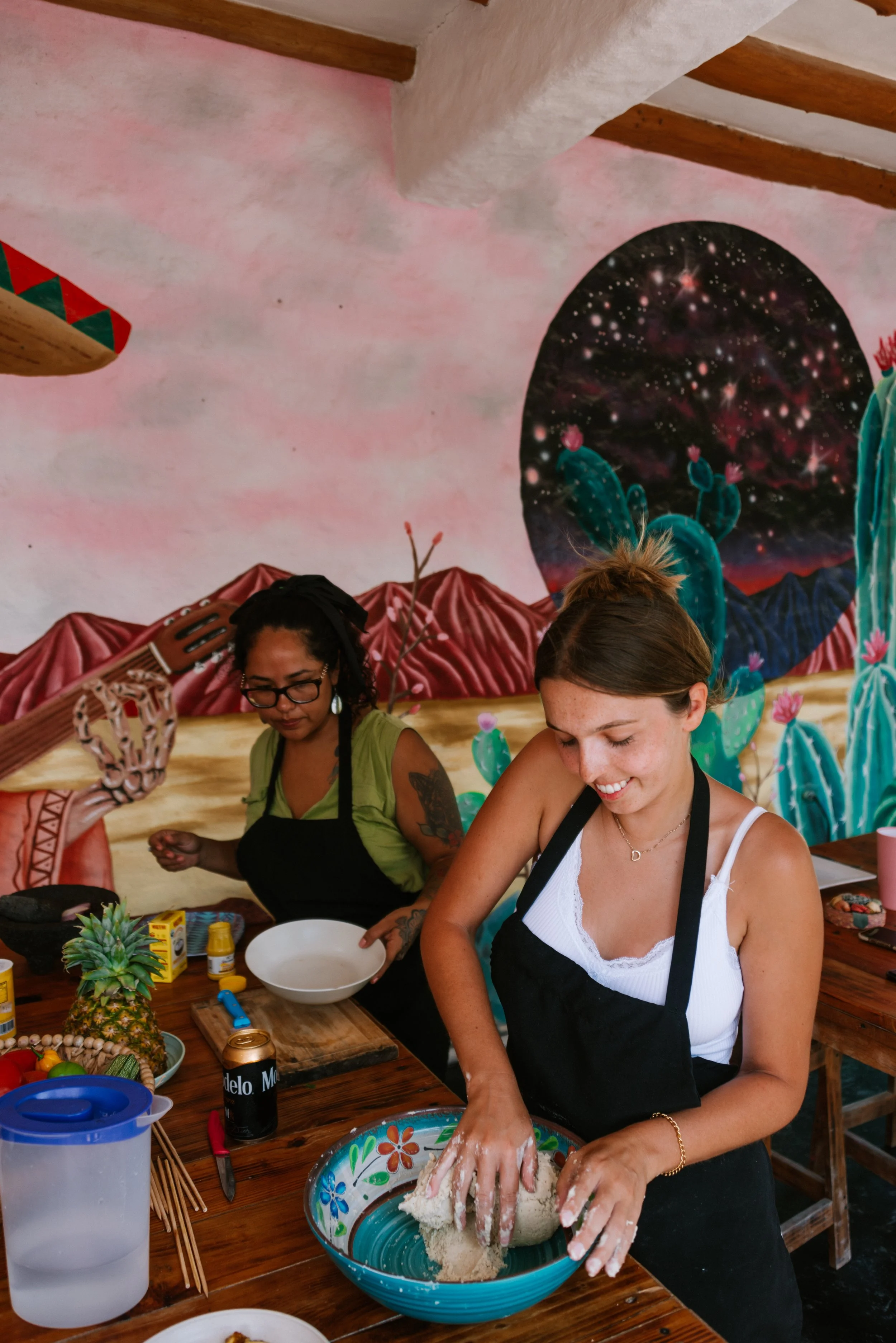 Two women cooking in a colorful, artistic kitchen with a mural of a galaxy and cactus plants on the wall.