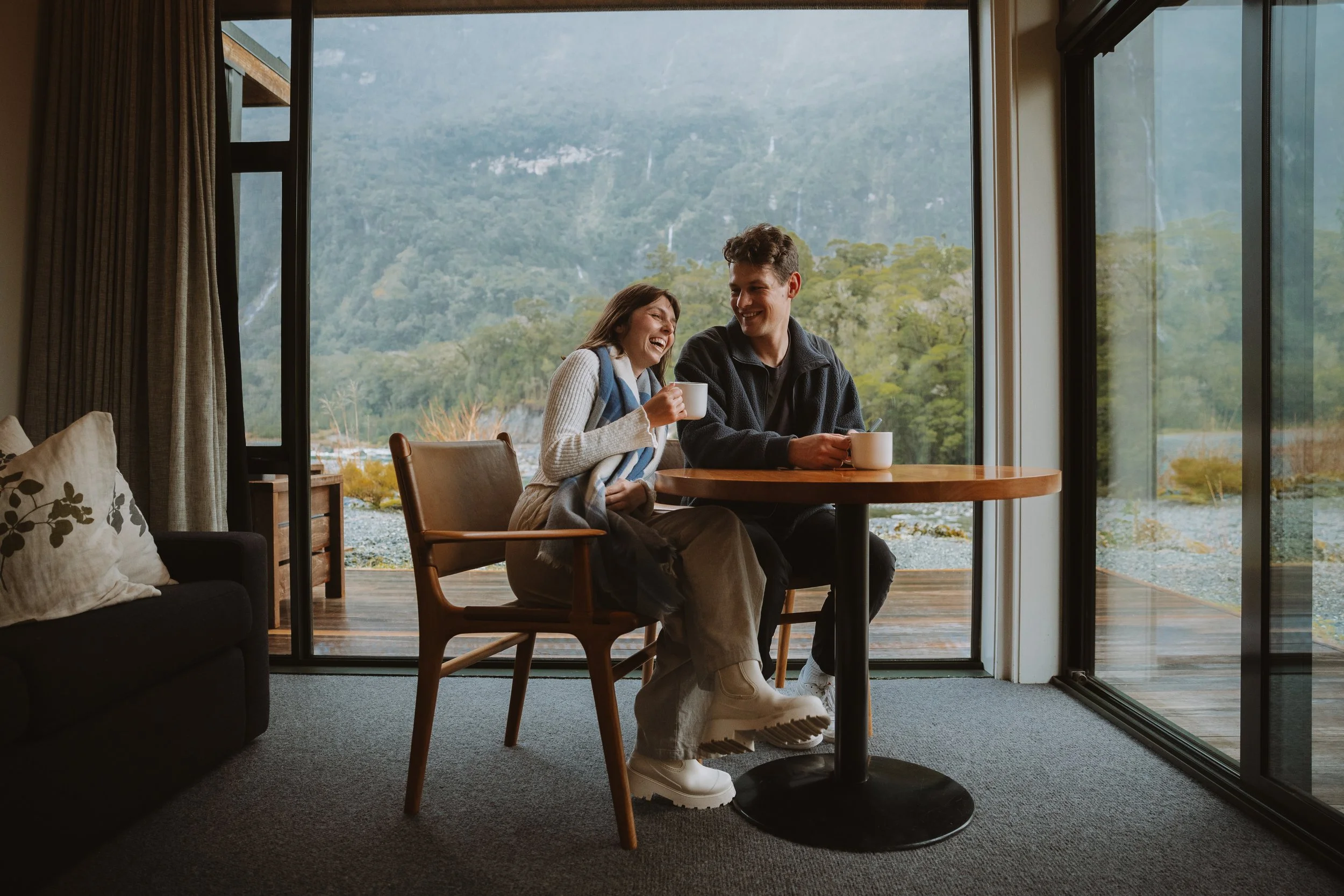 A man and woman sitting at a wooden table near large glass doors, enjoying coffee and laughing, with a mountain and forest view outside.