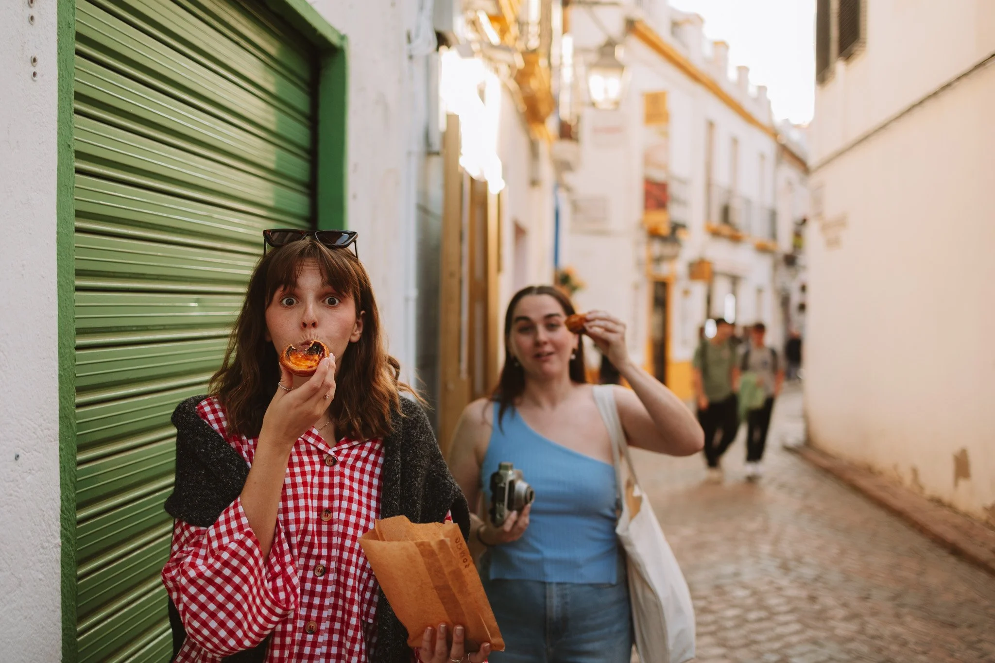 Two women are standing in a cobblestone street, eating pizza. The woman in the foreground has shoulder-length brunette hair, is wearing a red and white checkered shirt, and is holding a slice of pizza and a paper bag. The woman in the background has 